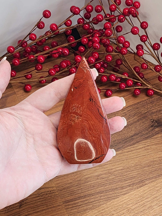A hand holding a red, triangular-shaped stone or gemstone, with a background of red berries and branches.