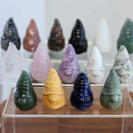 Adorable Crystal Gnomes around 1.9 inches tall on a clear display shelf in many materials including Goldstone, Green Aventurine, Sodalite, Green Jade, Clear Quartz, Rose Quartz, White Jade, Howlite, Peace Jade, and Lepidolite.