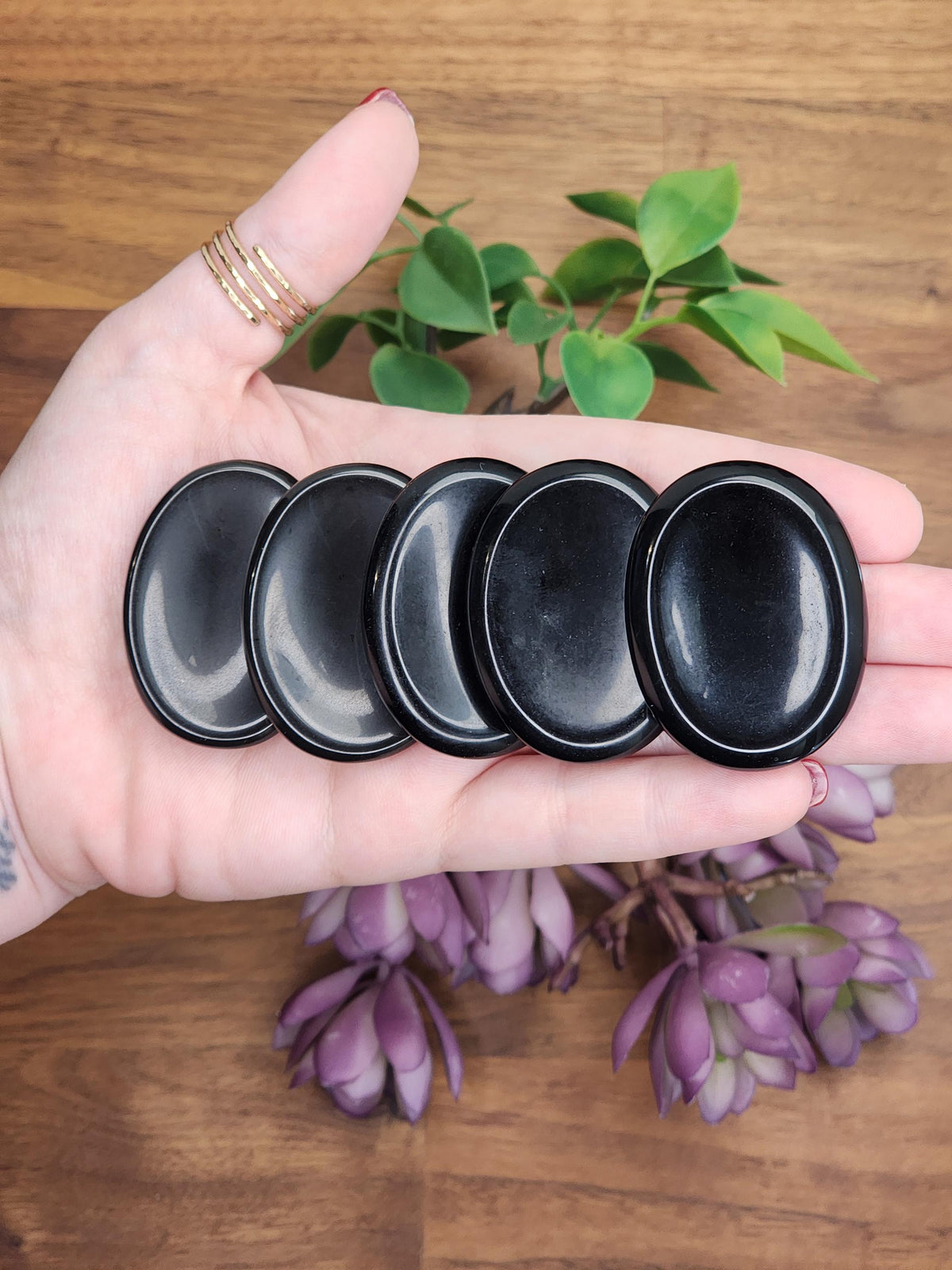 Black Obsidian Crystal Worry Stones sitting in palm of a caucasian hand with gold rings on the thumb. The 5 worry stones are oval shaped and a deep black color with highly polished finish reflecting the light of the camera.