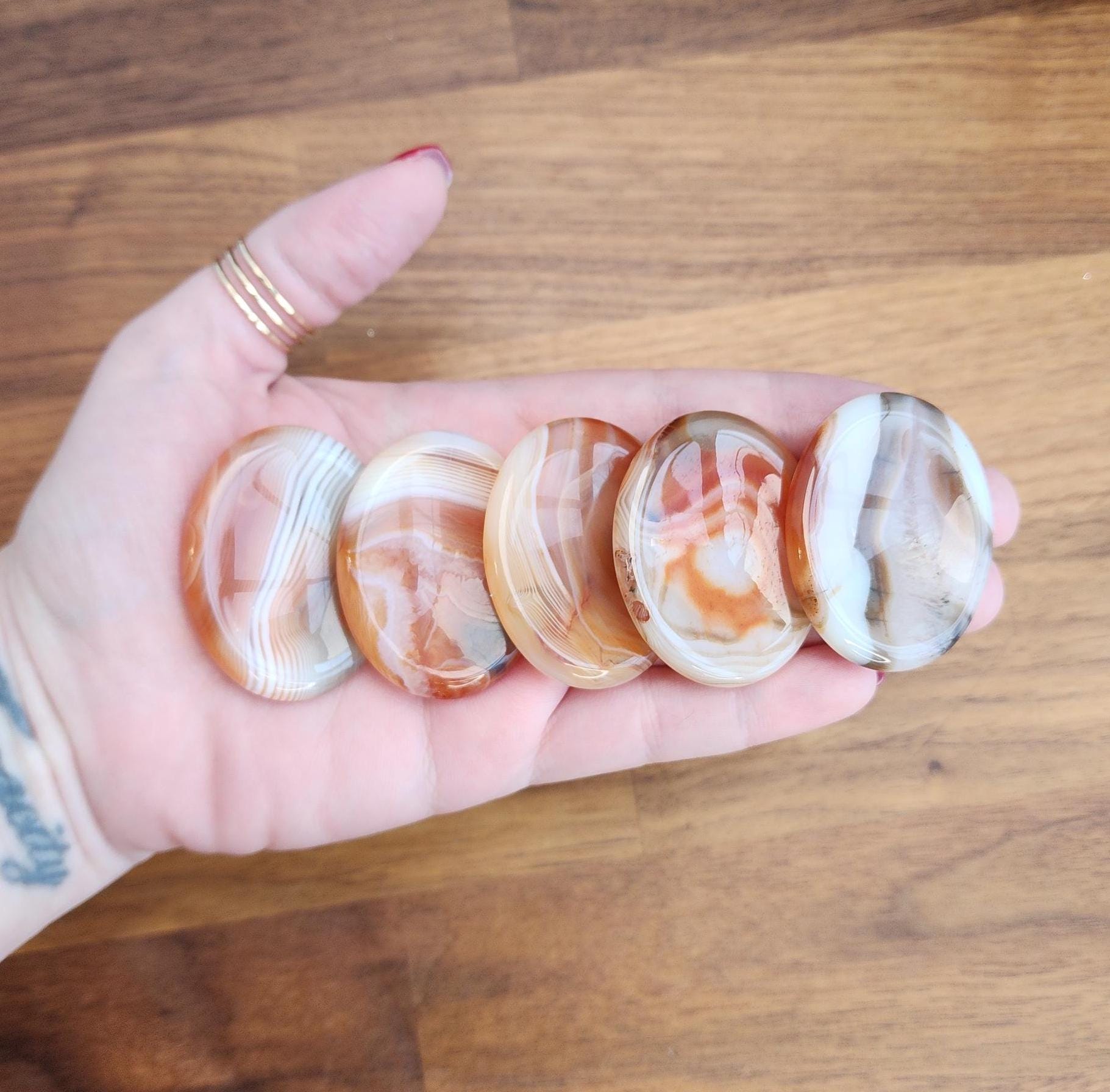 Crystal Worry stones in banded agate material on hand with gold rings on thumb above wood table top. The banded agate crystal worry stones are in tan, beige, red, orange, white and gray banding with beautiful patterns and swirls.