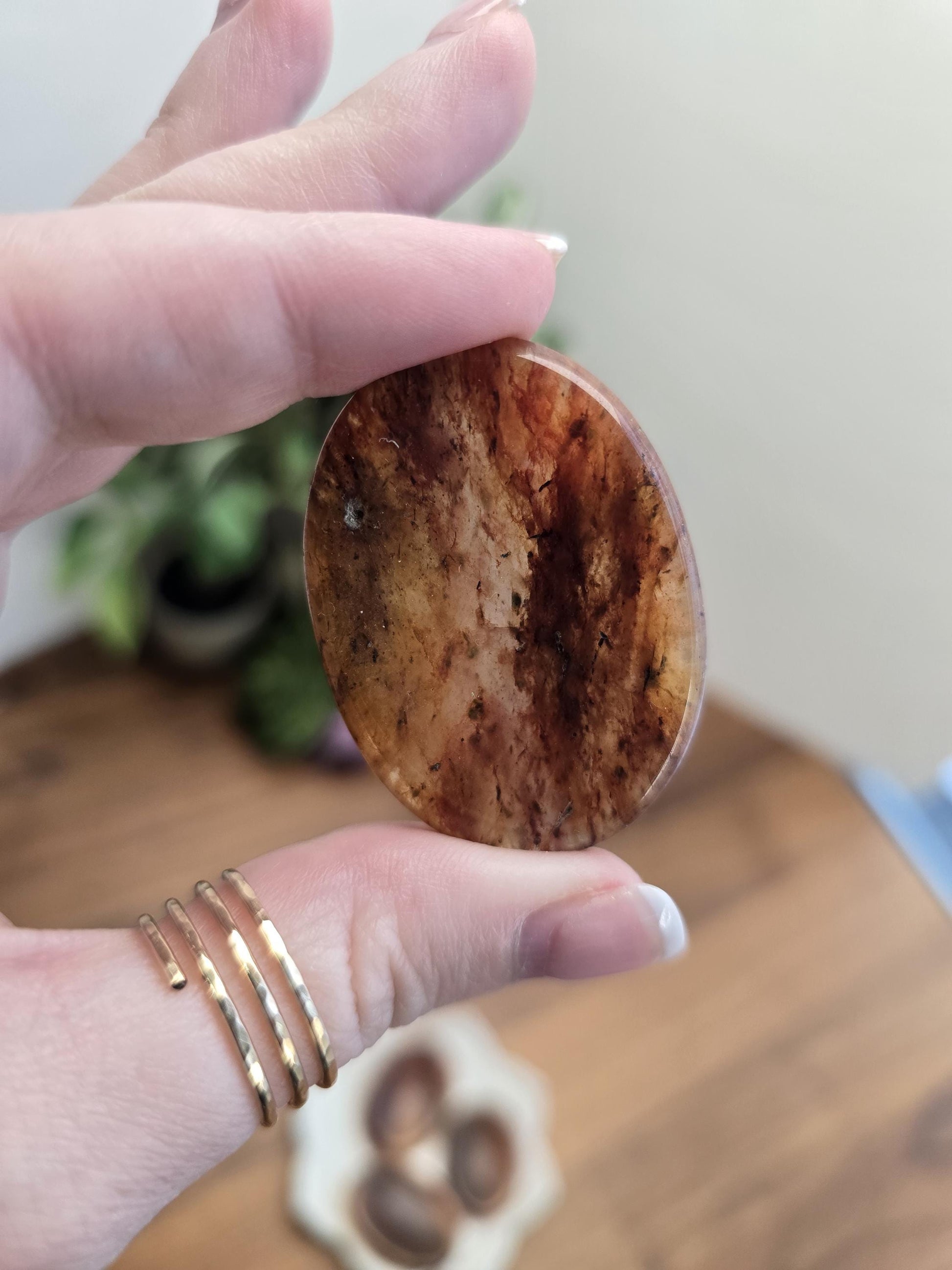 Gorgeous Hematoid Crystal Worry Stone with fiery patterns of Orange Red Brown and Black on a transparent Quartz matrix. Held in a finger and thumb aloft a wooden table