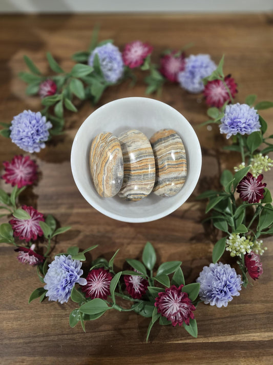 Three striped, oval-shaped, polished crystal stones in a white bowl. The stones are a mix of brown, yellow, and white caramel aragonite. The bowl is surrounded by a garland of purple, pink and green flowers.