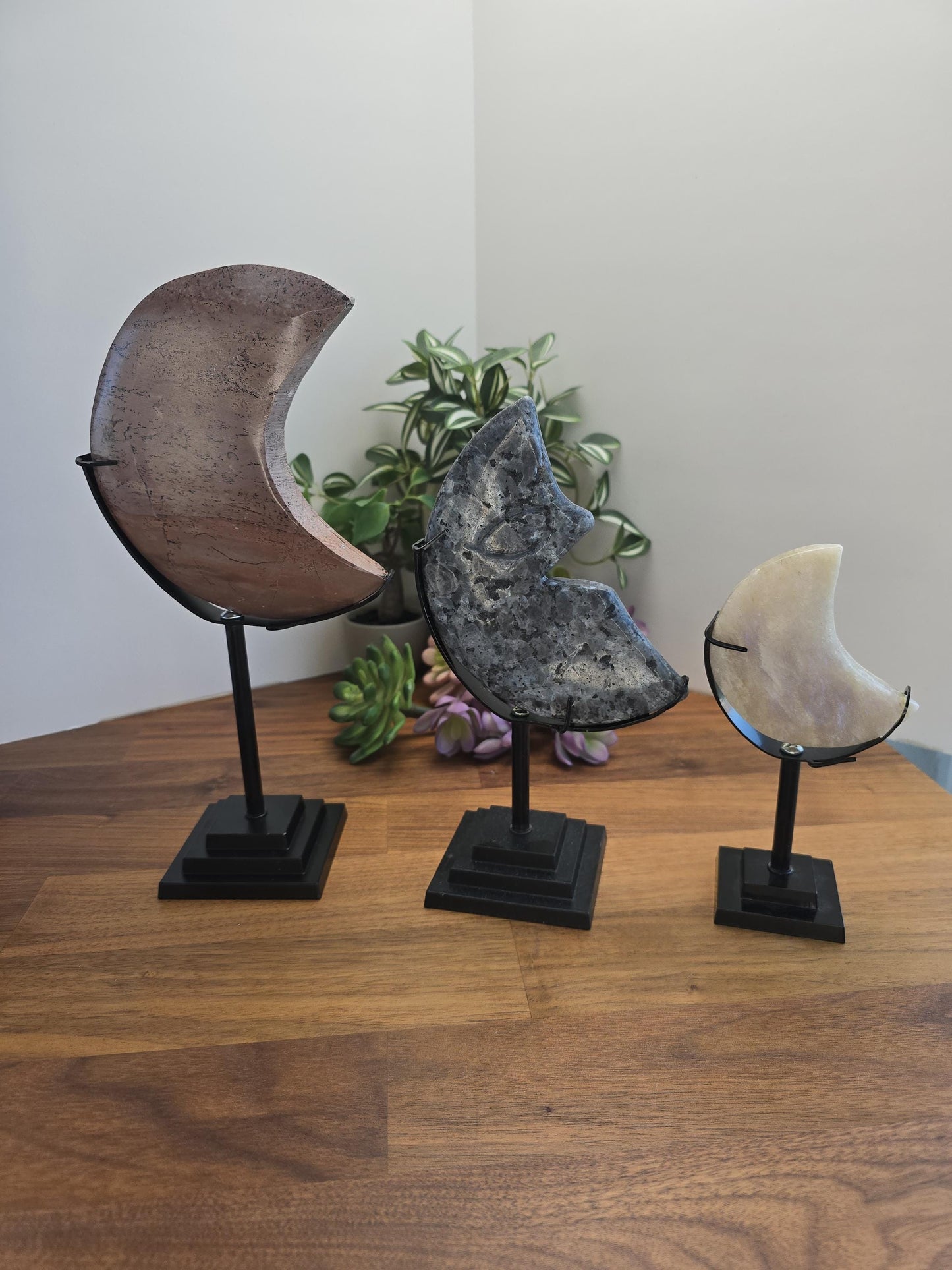 Large Medium and Small size black crystal moon stands. 3 black metal stands holding natural carved crystal moons on bendable stands standing on wood desk.