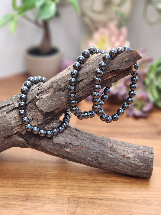 May include: A black hematite beaded bracelet with a shiny finish. The bracelet is wrapped around a piece of driftwood.