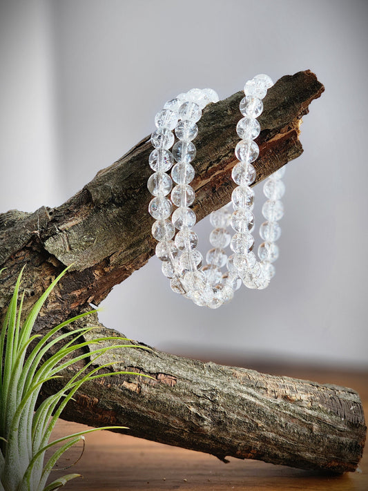 May include: A strand of clear quartz crystal beads, with a faceted finish, draped over a piece of driftwood.
