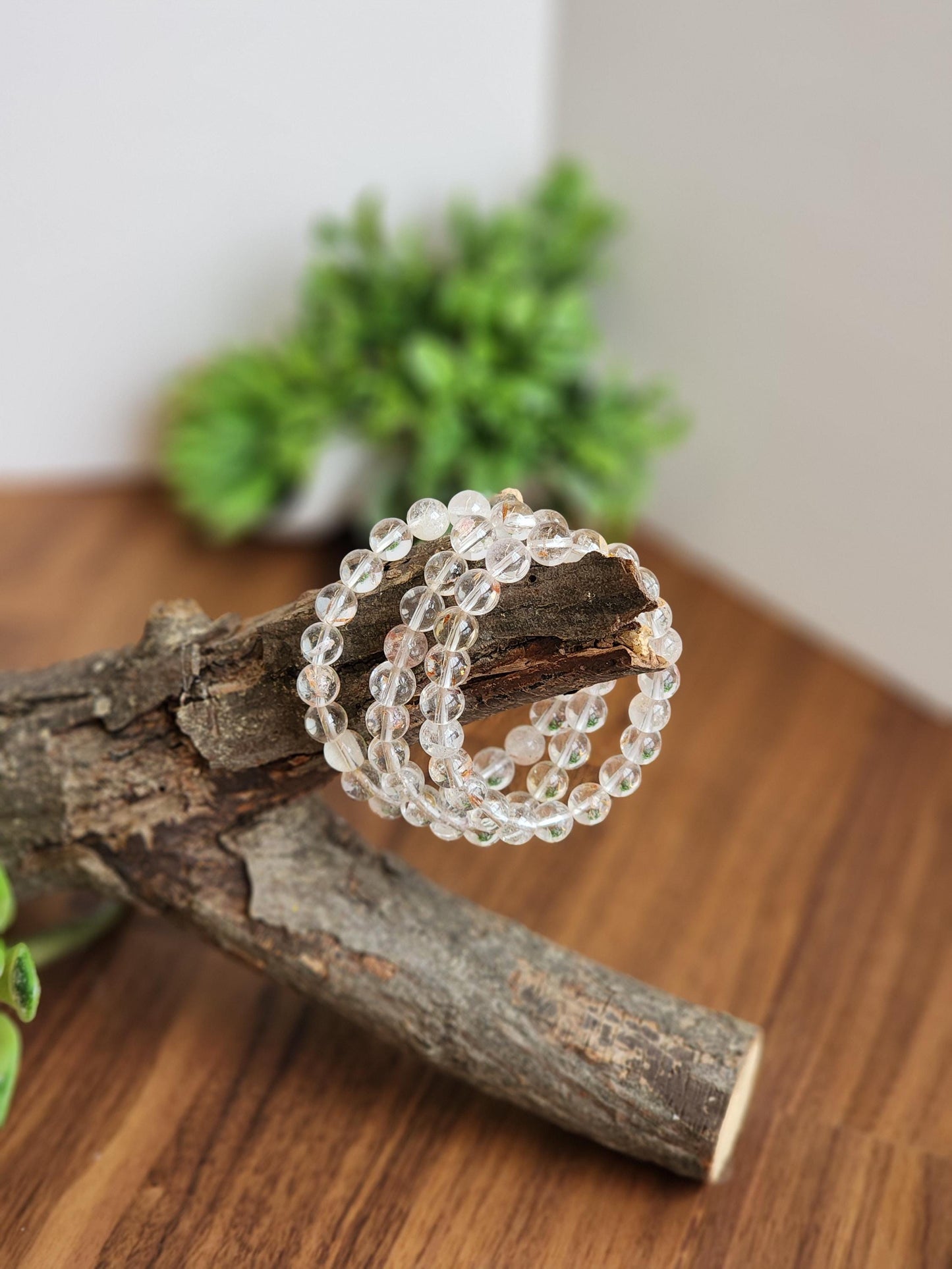May include: Clear quartz crystal bead bracelets, displayed on a piece of wood. The bracelets are made of round, translucent beads. The background includes a blurred green plant and a wooden surface.