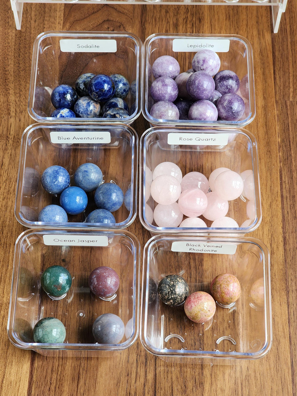 six clear plastic containers filled with various colored gemstones, including blue, purple, pink, and green stones, arranged on a wooden surface.