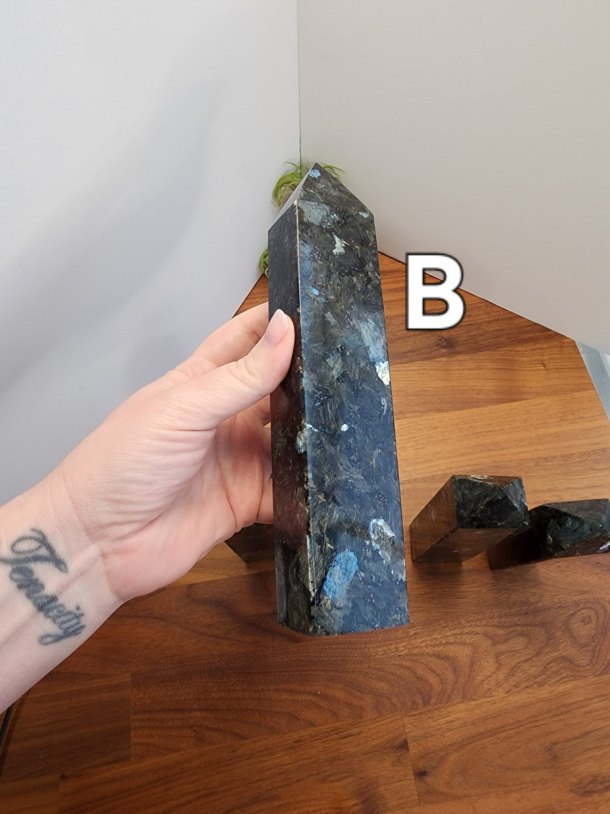 May include: A dark blue and green labradorite crystal point with a pointed tip. The crystal is about 6 inches long and is sitting on a wooden surface.