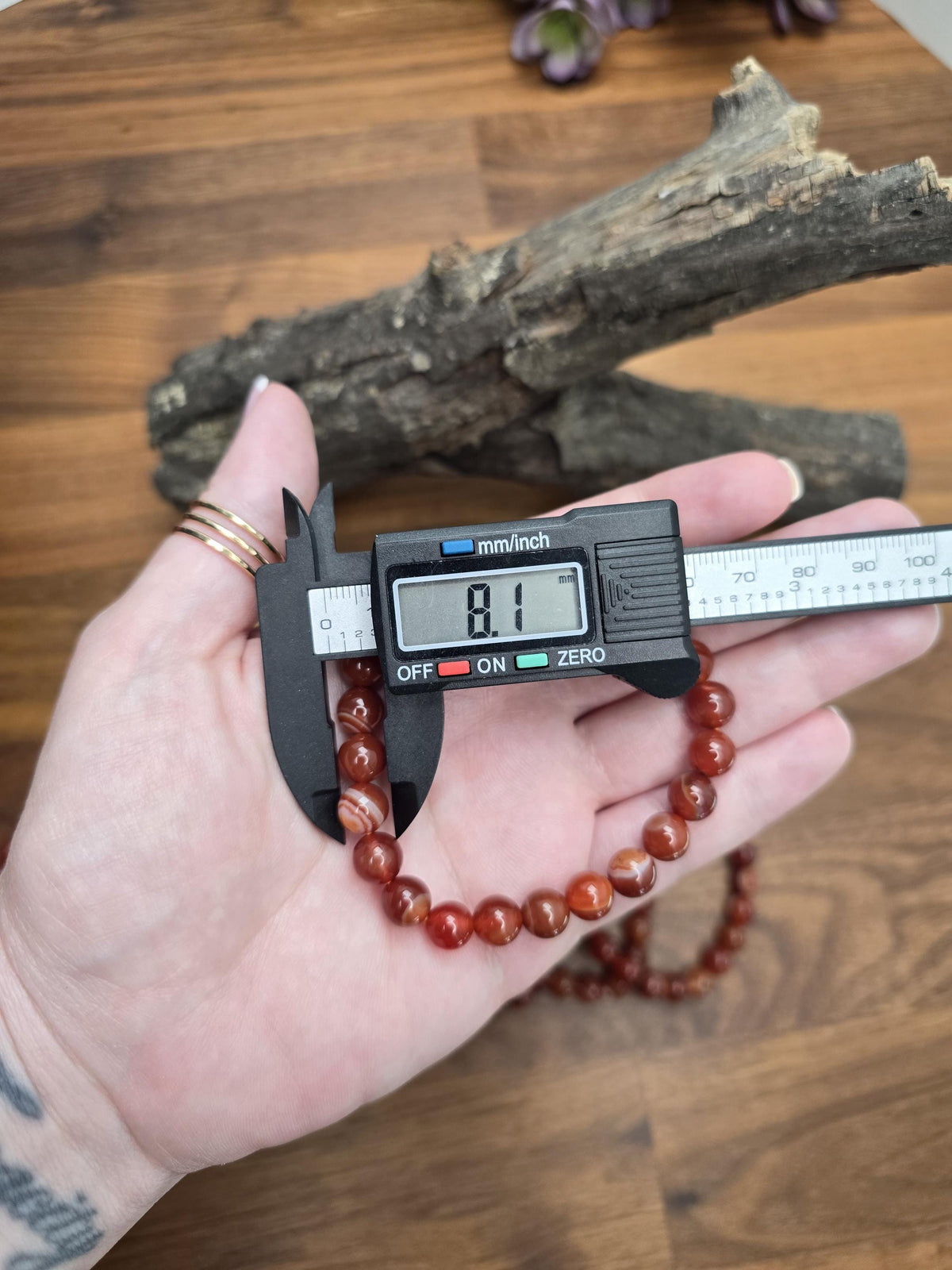 A person's hand holding a digital caliper to measure the length of a red bead bracelet.