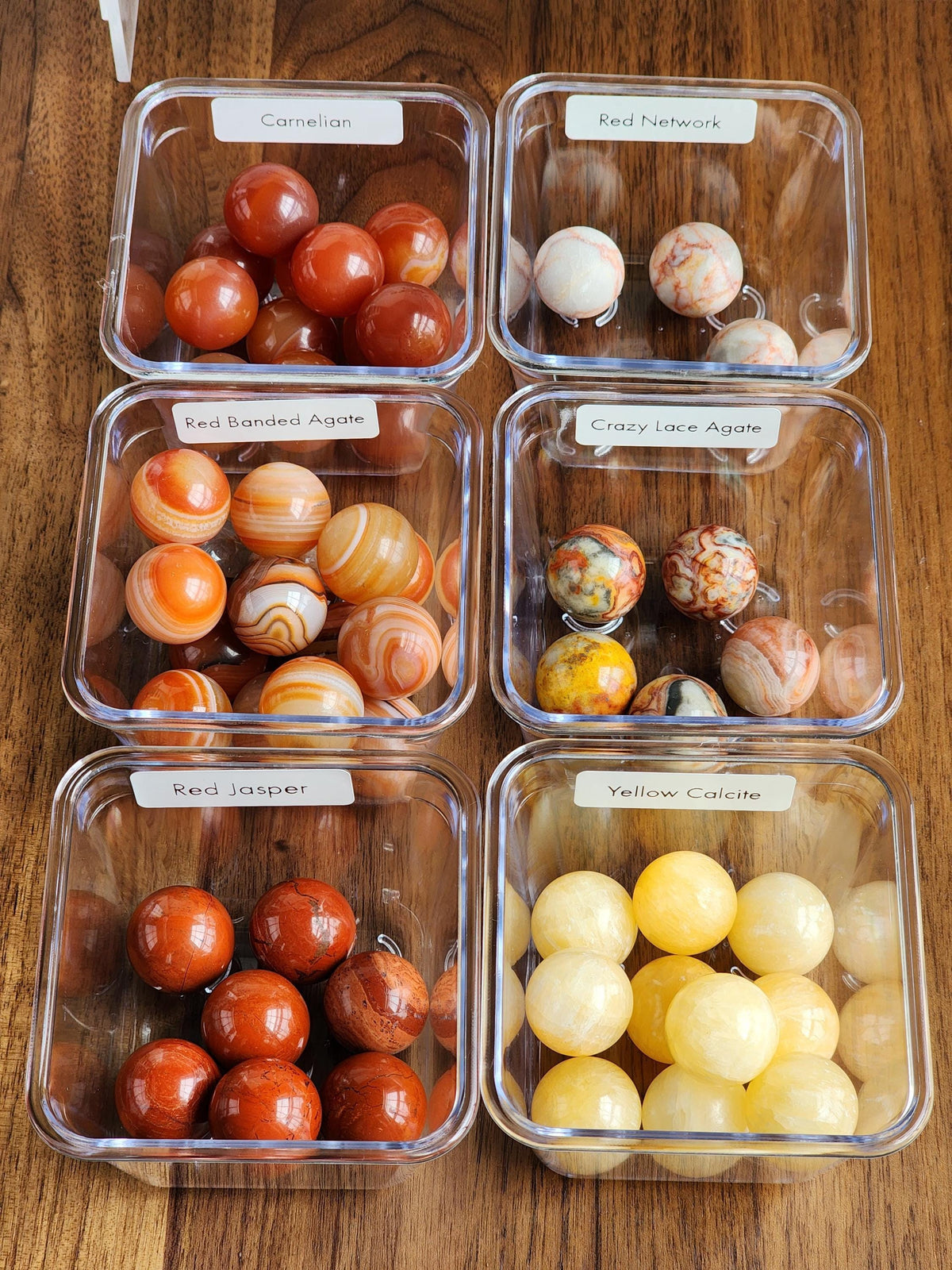 six clear plastic containers filled with various colored stones, including red, yellow, and orange stones, arranged on a wooden surface.