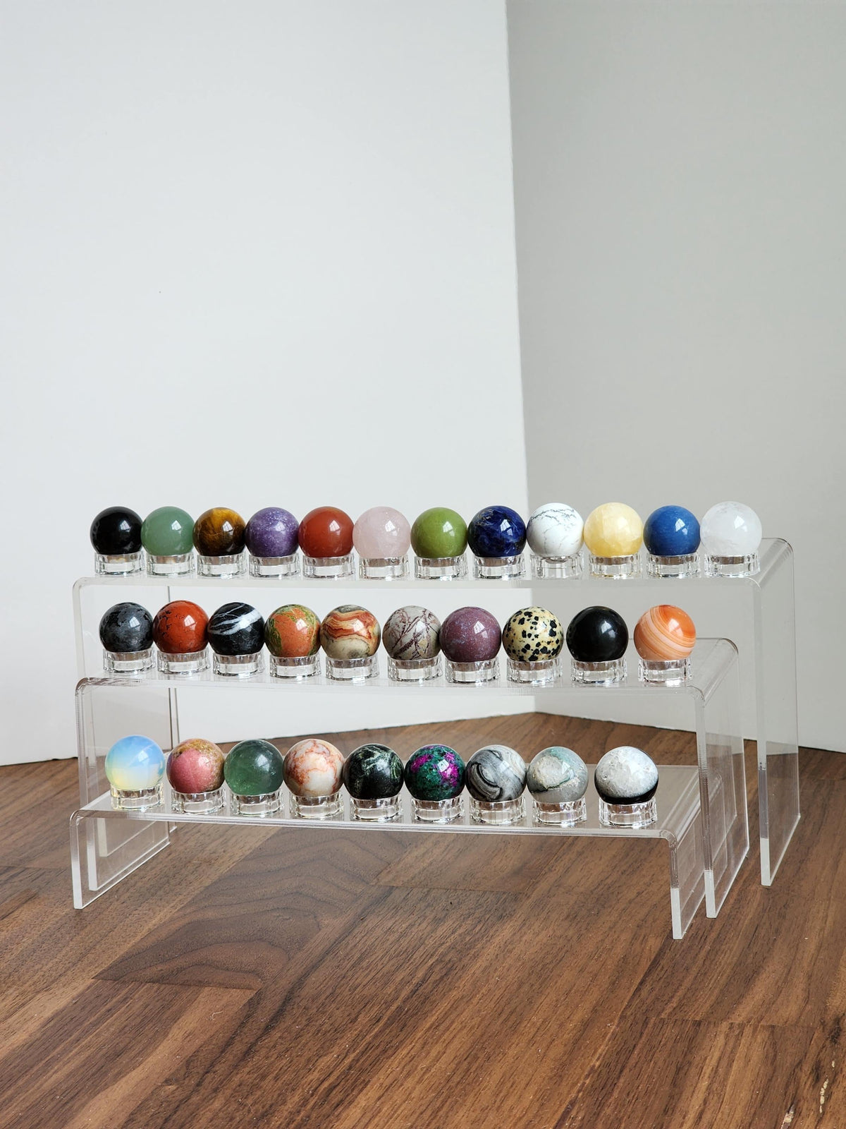 A display of various colorful gemstones and minerals arranged on three clear glass shelves against a white wall.