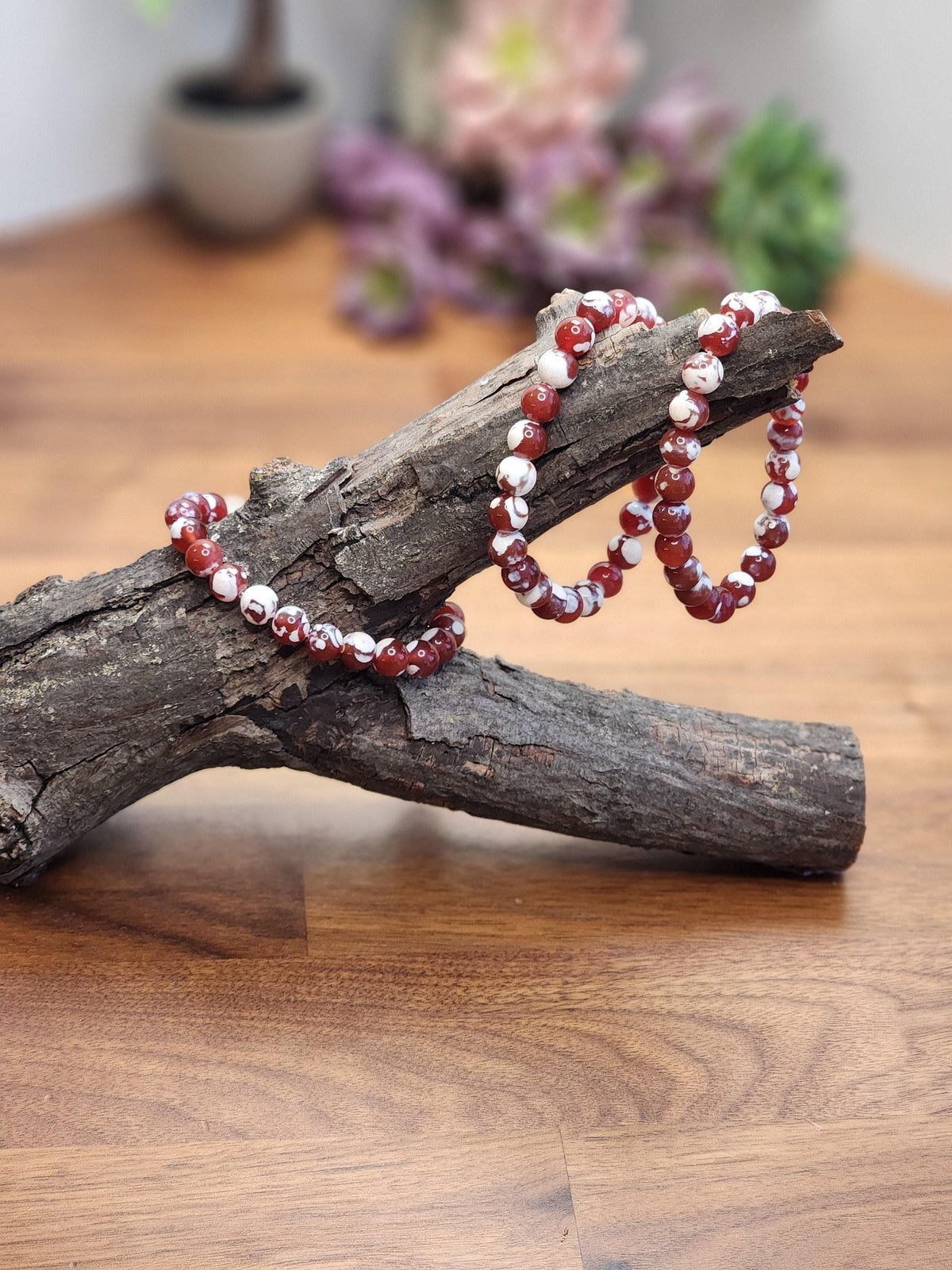 a pair of red and white beaded bracelets resting on a wooden surface, with a blurred background of flowers and foliage.