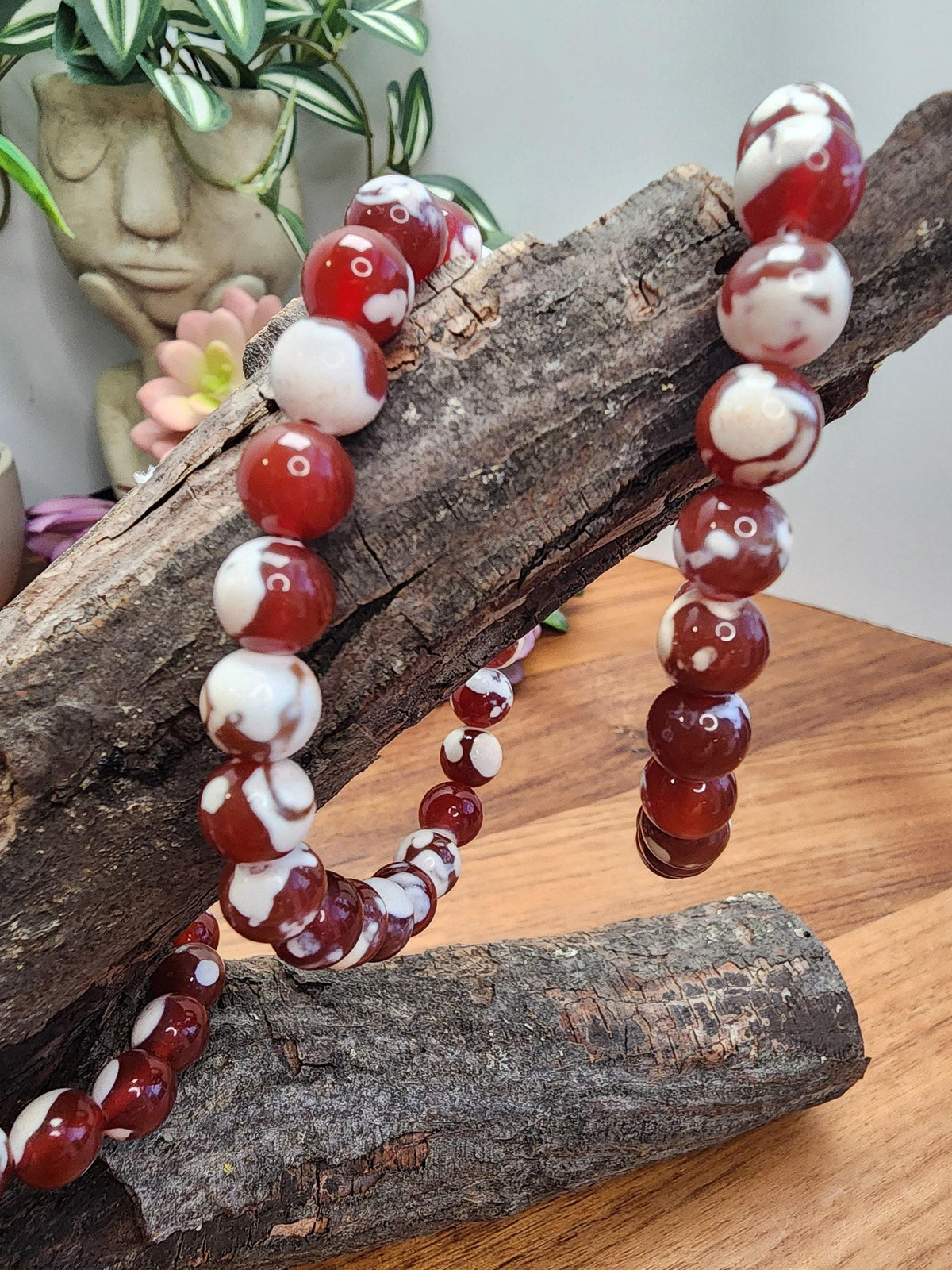 A necklace of red and white beads is displayed on a wooden surface, with a potted plant and a vase visible in the background.