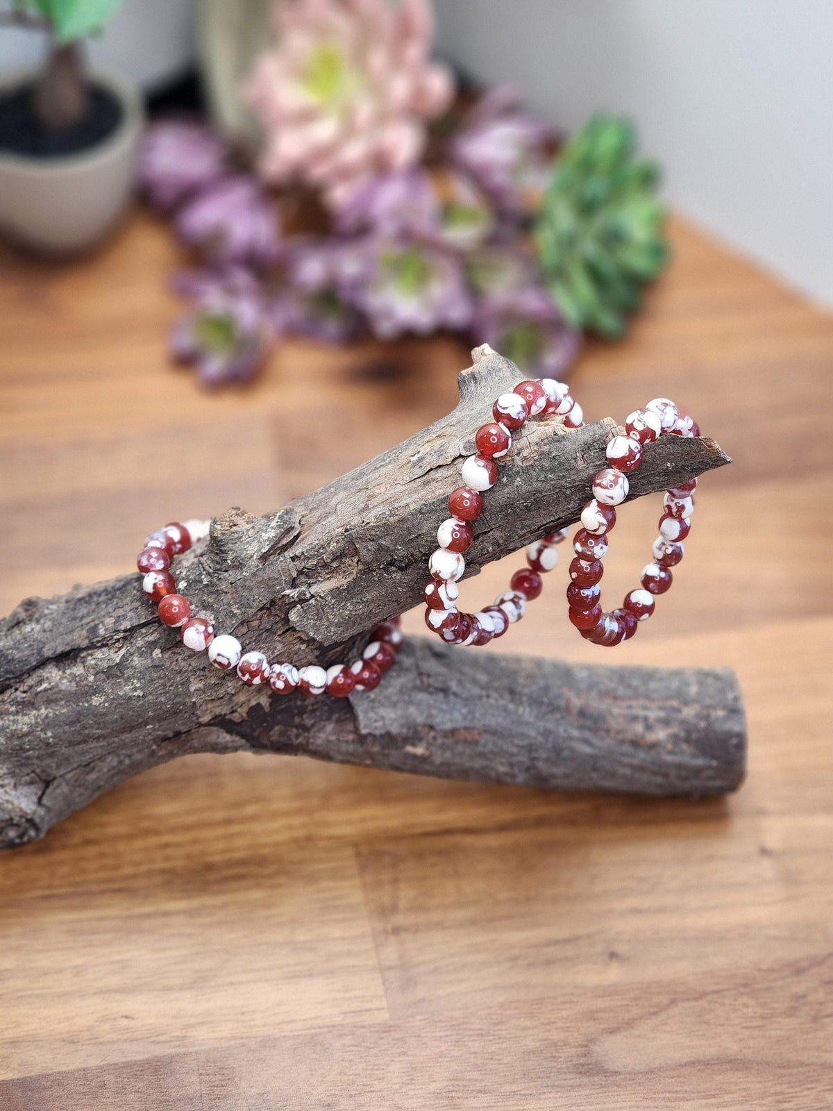 two red and white beaded bracelets resting on a wooden surface, with a blurred background of flowers and foliage.