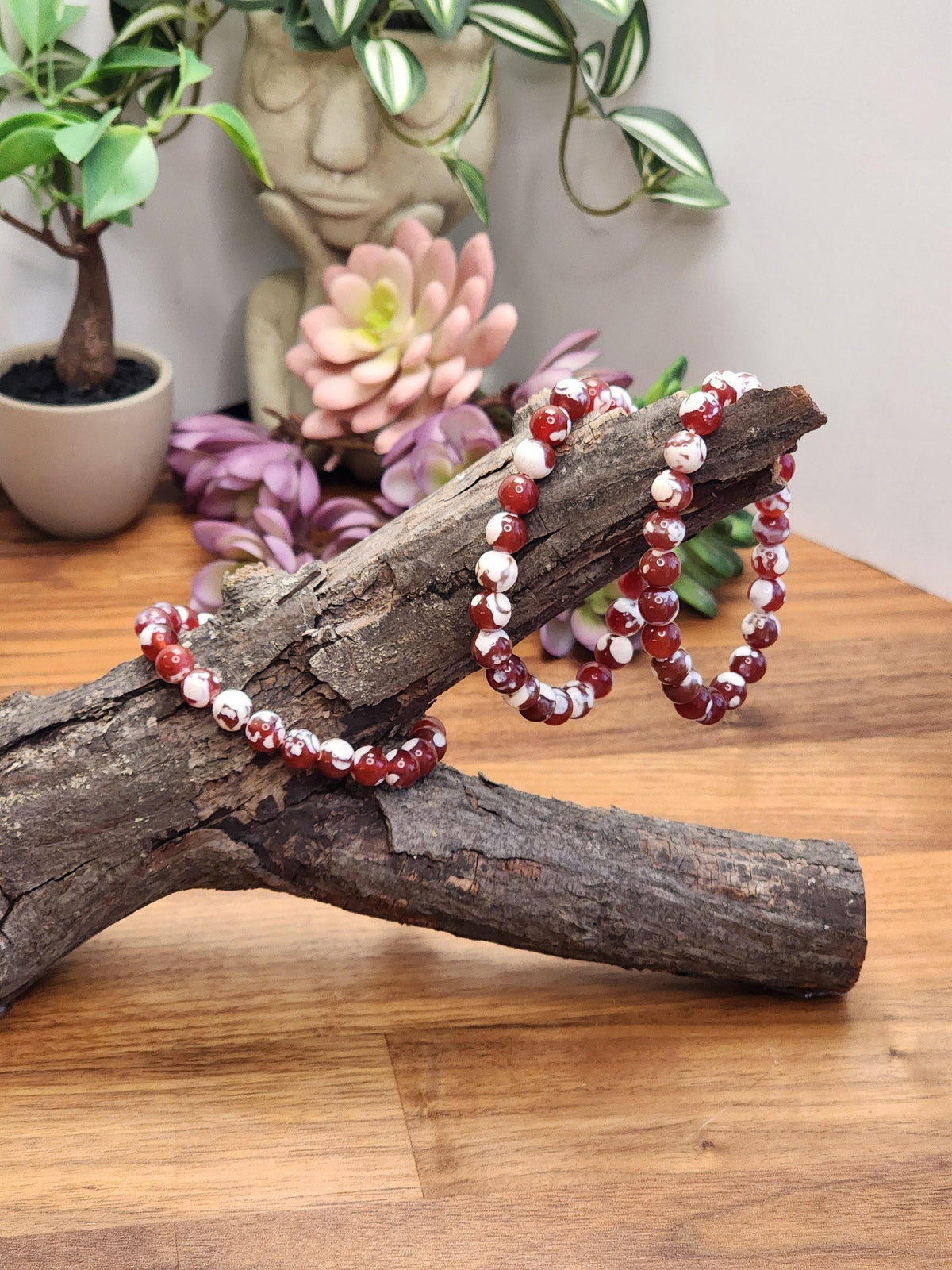 a wooden surface with two red and white beaded bracelets resting on a branch, surrounded by various potted plants and flowers.