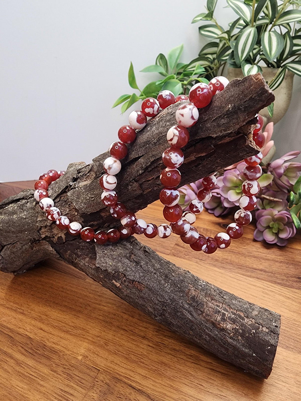 a pair of red and white beaded bracelets resting on a wooden surface, with a potted plant and flowers in the background.