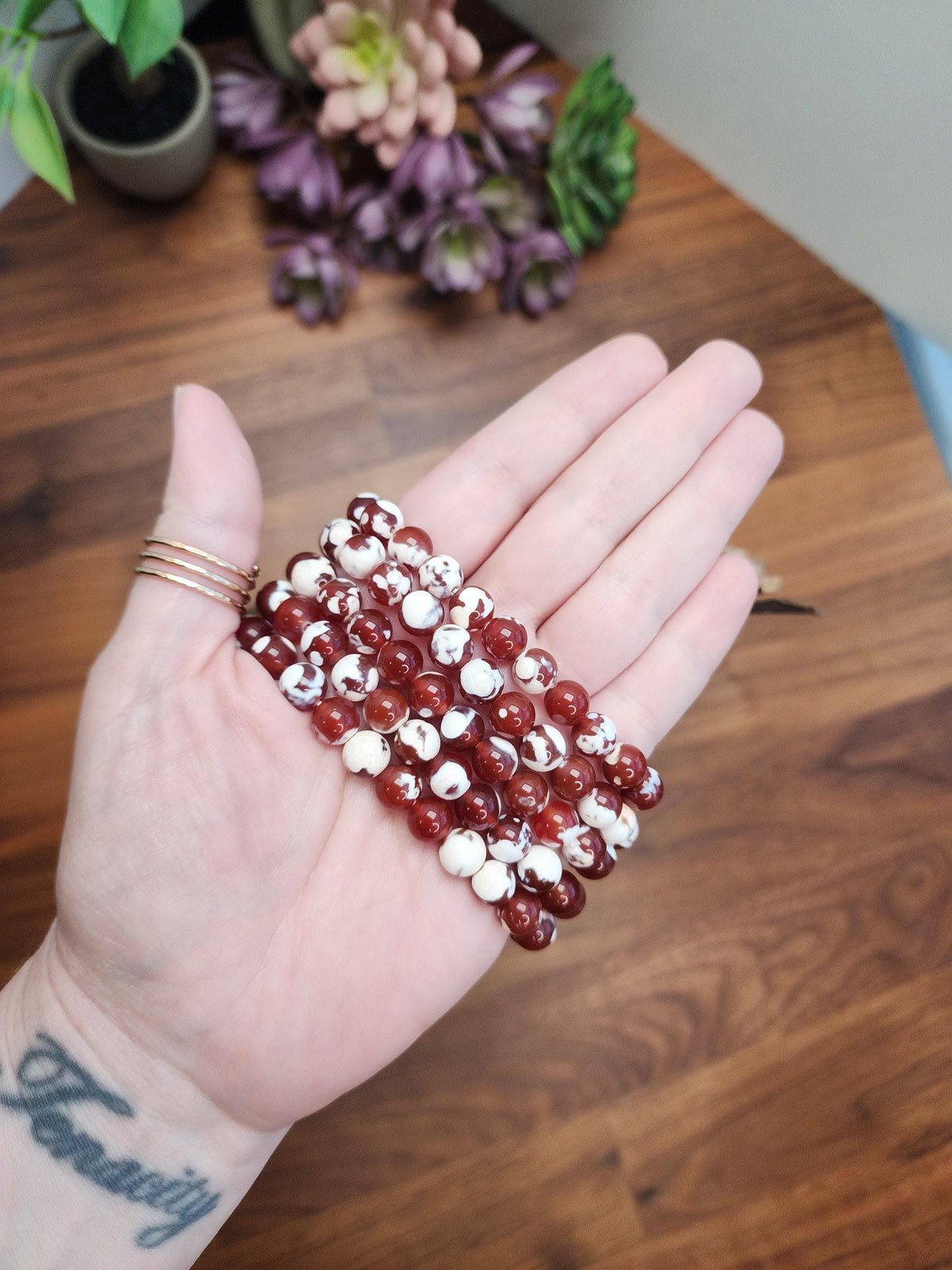 A hand holding a stack of red and white beads against a wooden background with purple flowers.