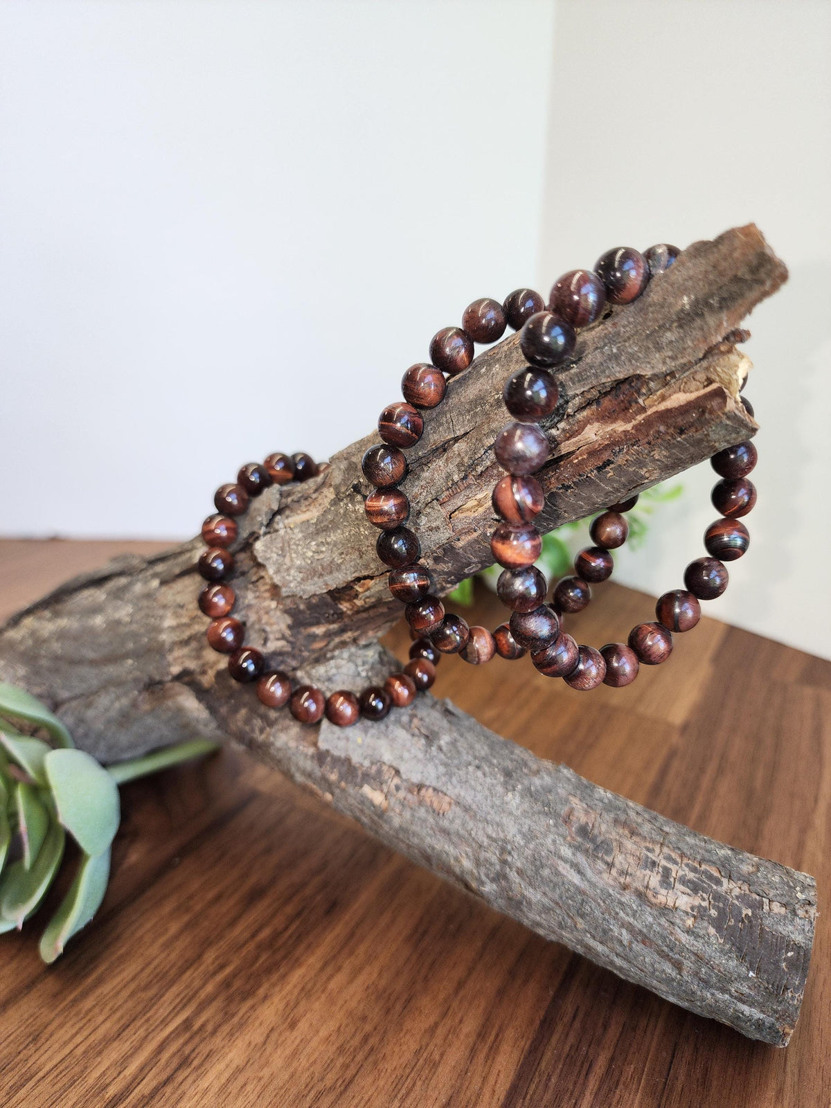 a pair of brown beaded bracelets resting on a wooden surface, with a plant leaf visible in the background.