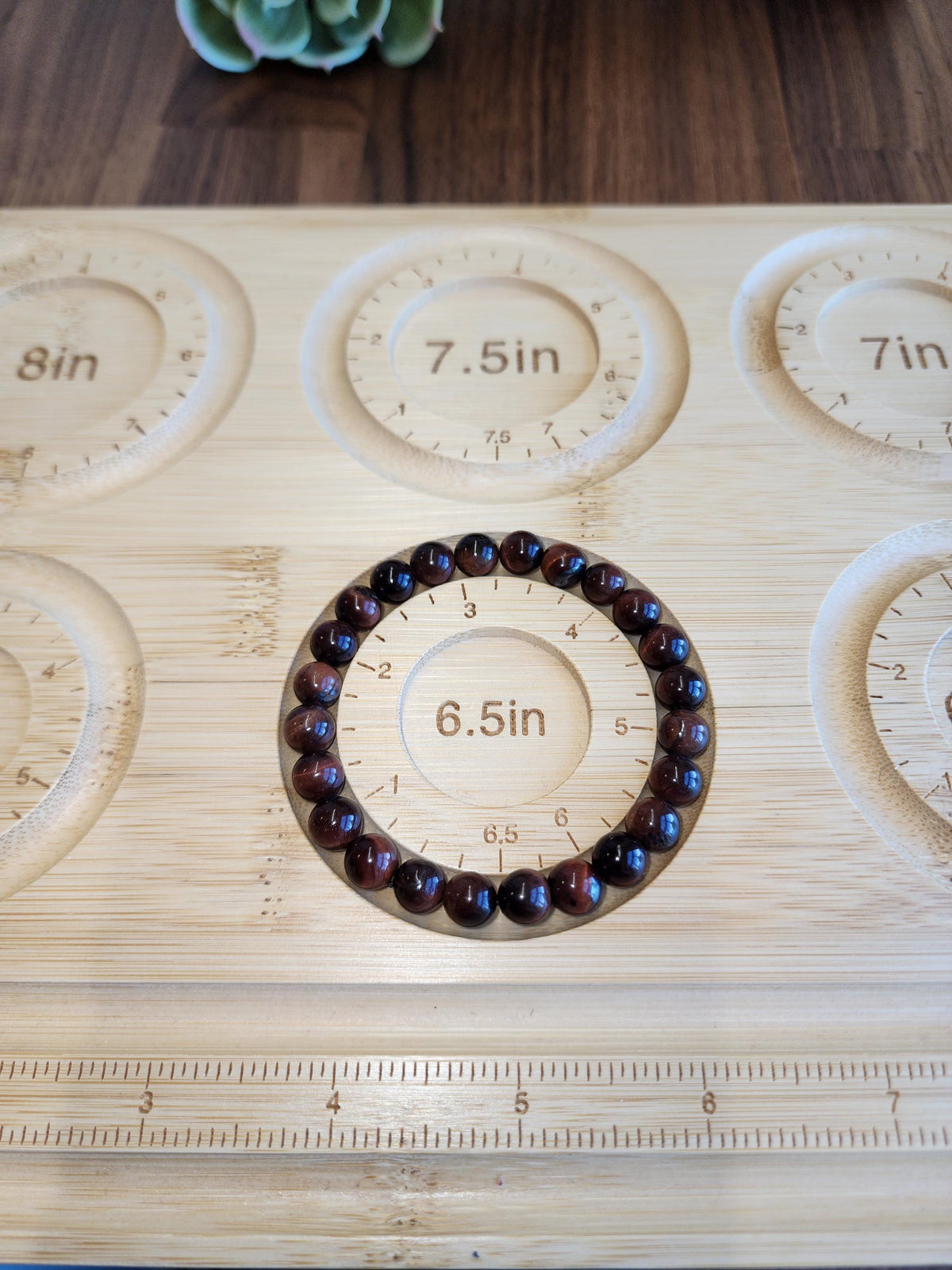 a wooden board with a bracelet and a ruler placed on it. The bracelet is made of beads and is positioned in the center of the board, surrounded by a ruler for size reference.