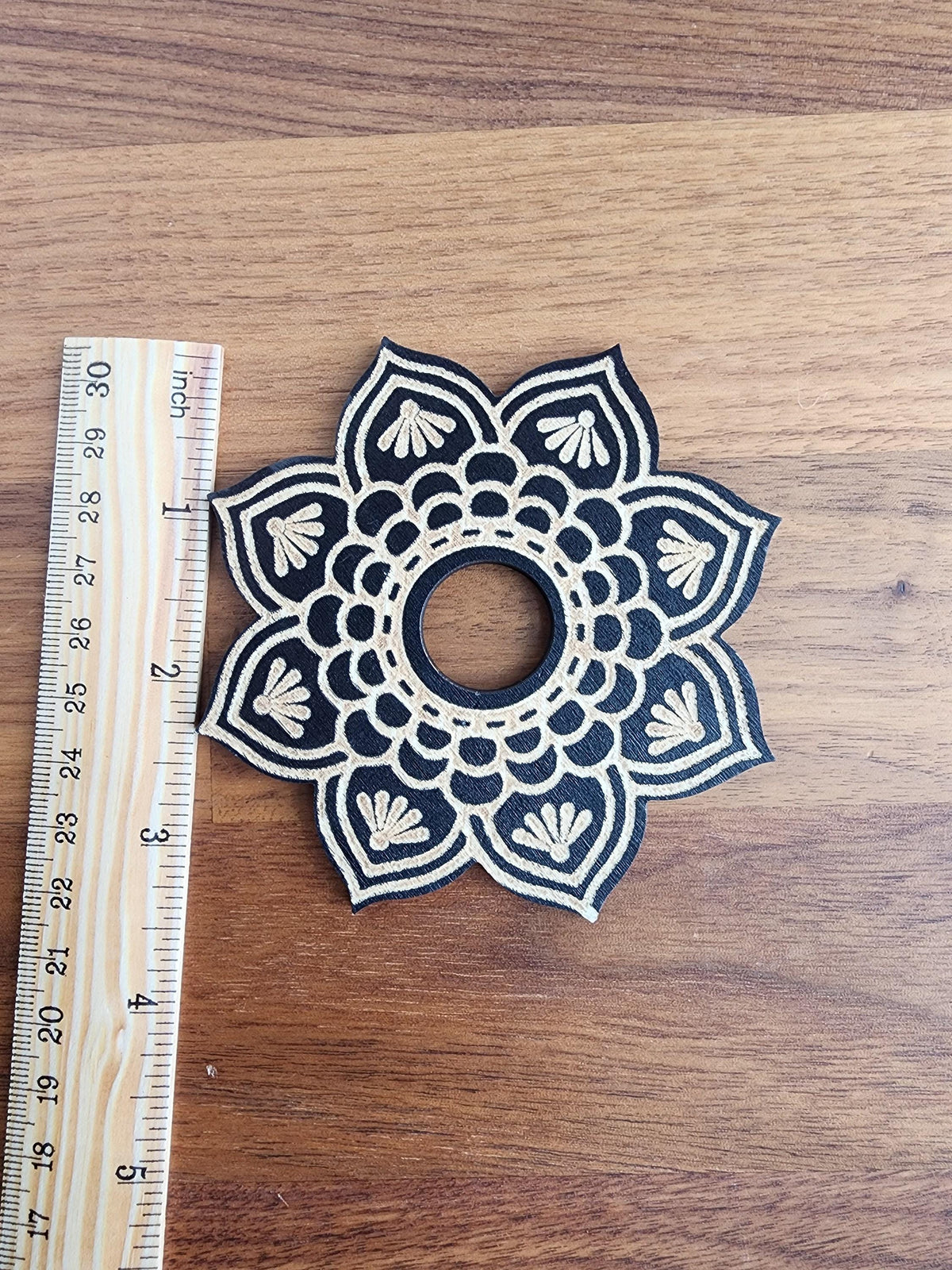 A black and white ornate flower-like design with a central hole is placed next to a ruler on a wooden surface.