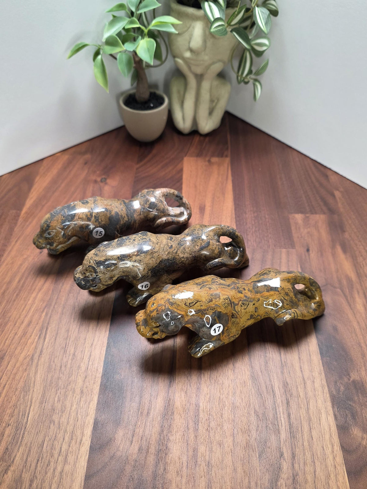 Three carved stone animal figurines are displayed on a wooden surface, with a potted plant and a vase in the background.