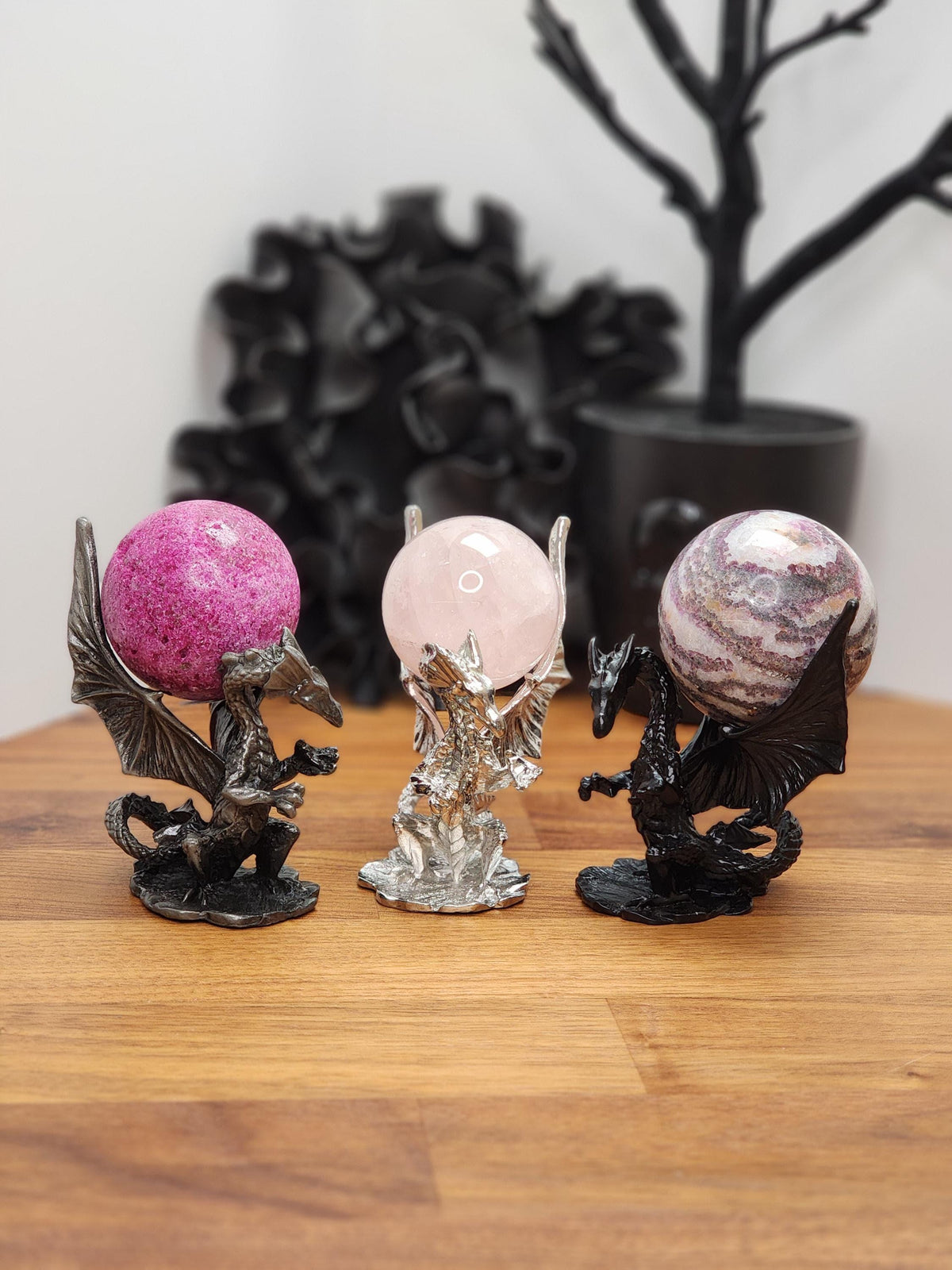 Three dragon figurines in different colors - pink, silver, and black - stand on a wooden surface, with a black plant in the background. Black dragon holds zebra fluorite crystal. Silver dragon holds rose quartz ball. Brushed silver color holds uv reactive honeycomb ruby