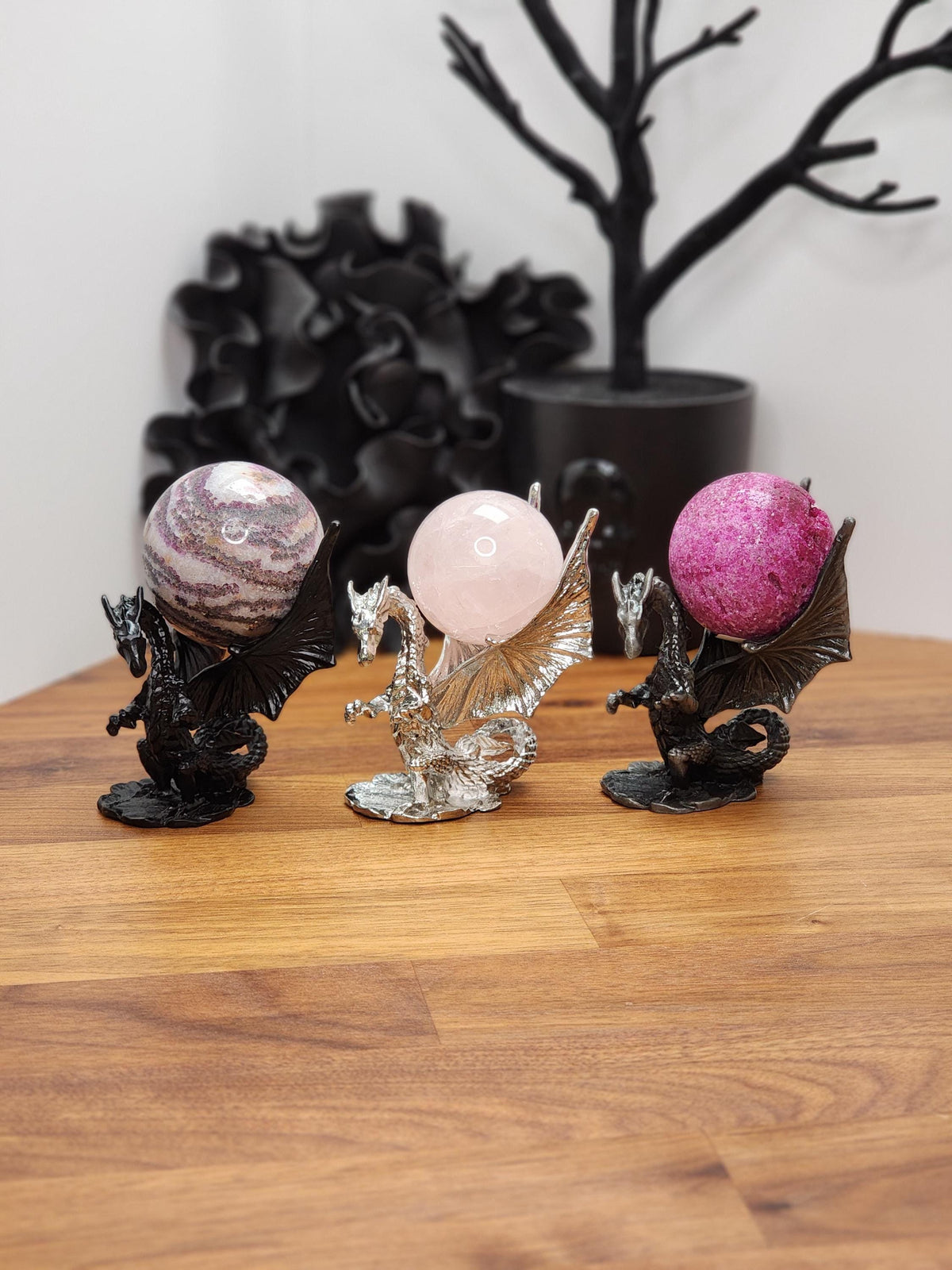 three dragon figurines on a wooden surface, each holding a different colored gemstone sphere. Black dragon holds zebra fluorite crystal. Silver dragon holds rose quartz ball. Brushed silver color holds uv reactive honeycomb ruby