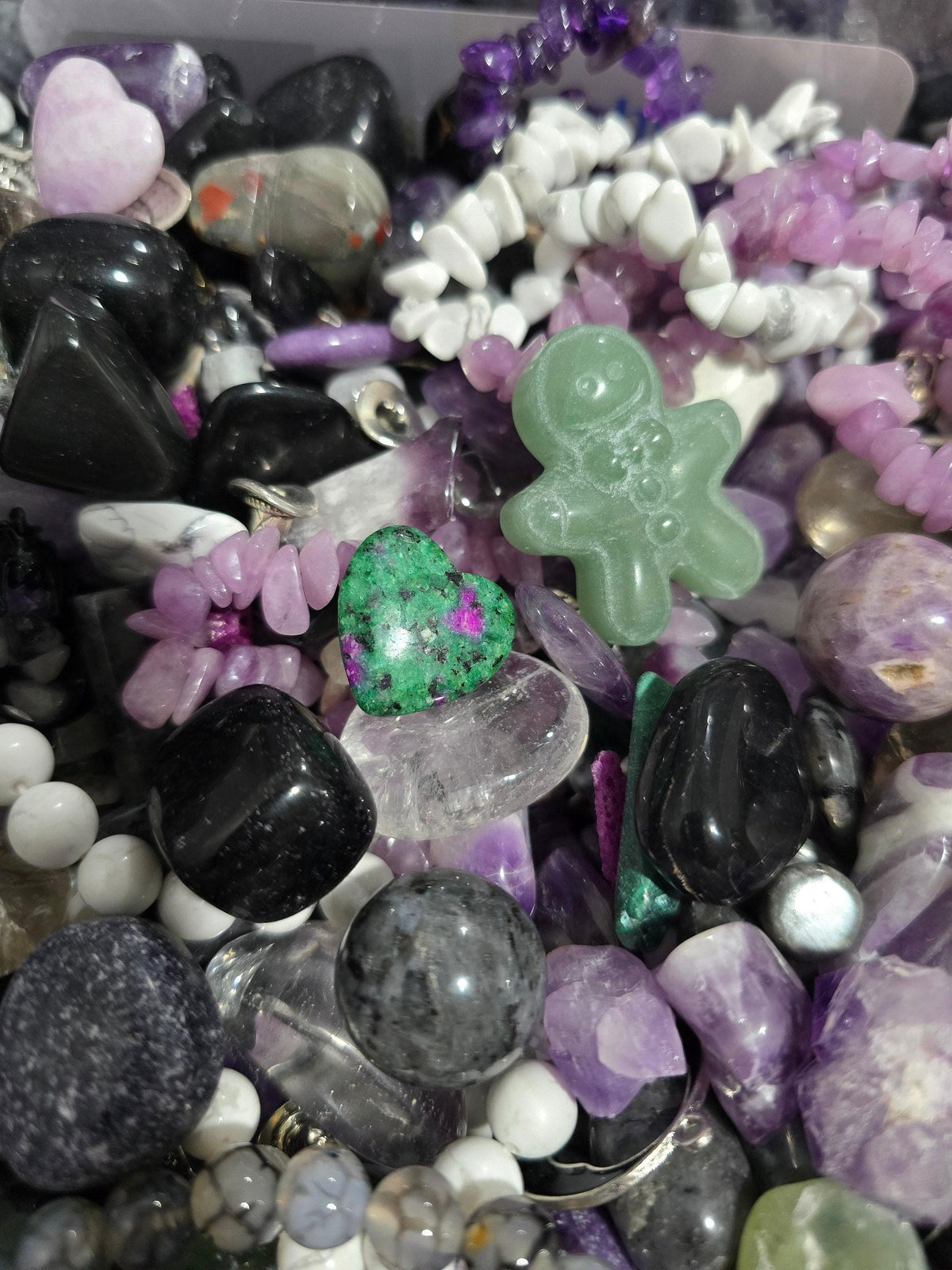 The image is a close-up of a collection of various gemstones and crystals, including amethyst, jade, and other colorful stones, as well as some beads and charms.