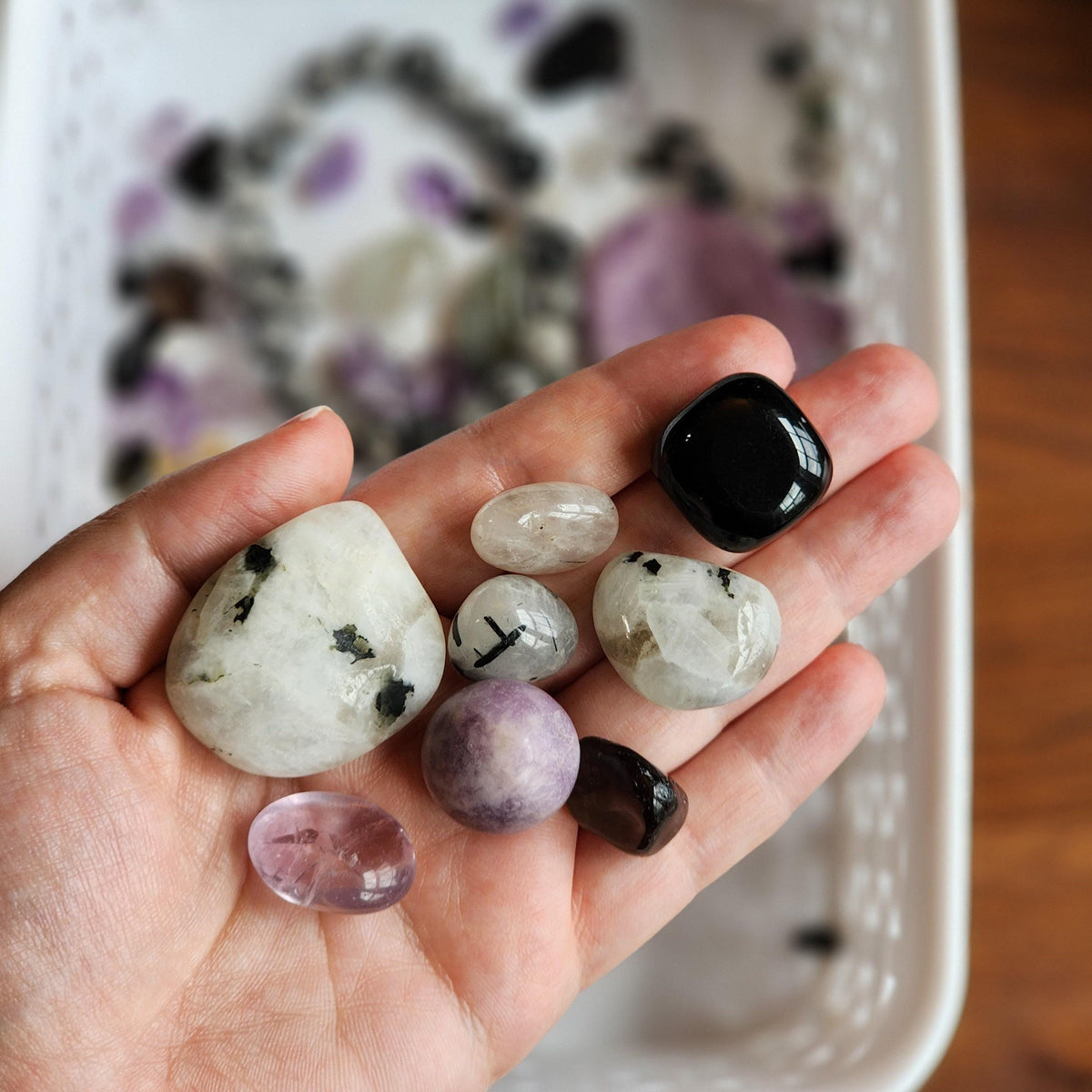 A hand holding various colored and shaped stones, including white, black, purple, and clear stones, against a blurred background.
