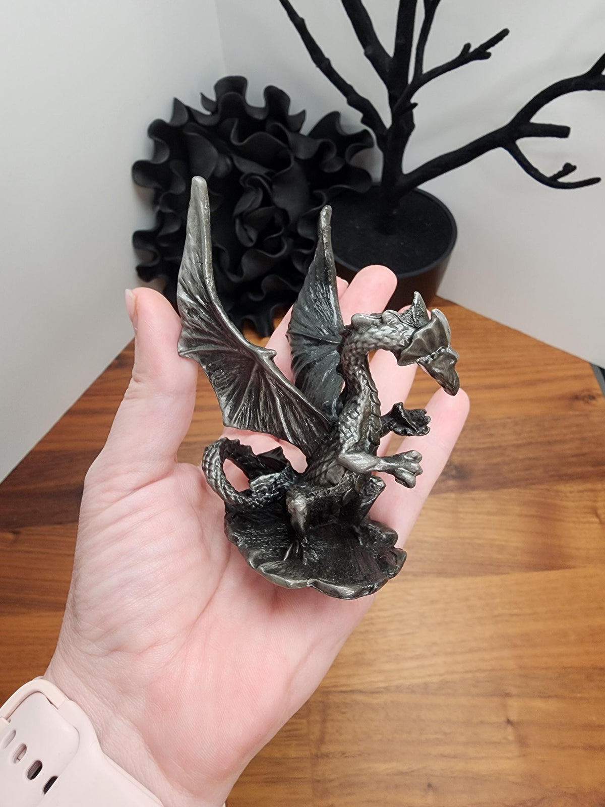 A person's hand is holding a small metal dragon figurine, which appears to be a dragon-like creature with wings and a tail. The background features a wooden surface and a black plant or sculpture.