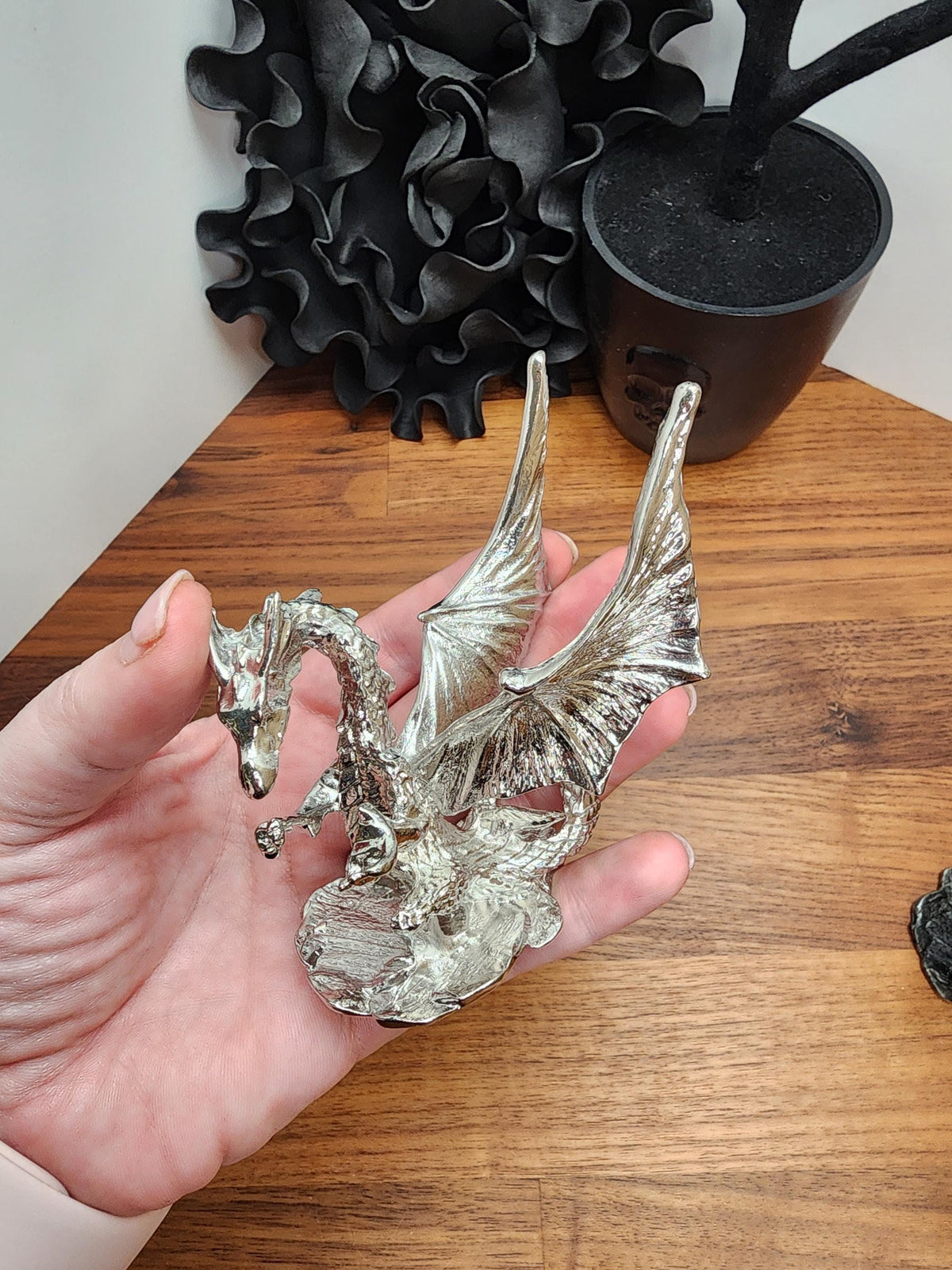 A hand is holding a silver dragon figurine with wings spread, against a wooden background.