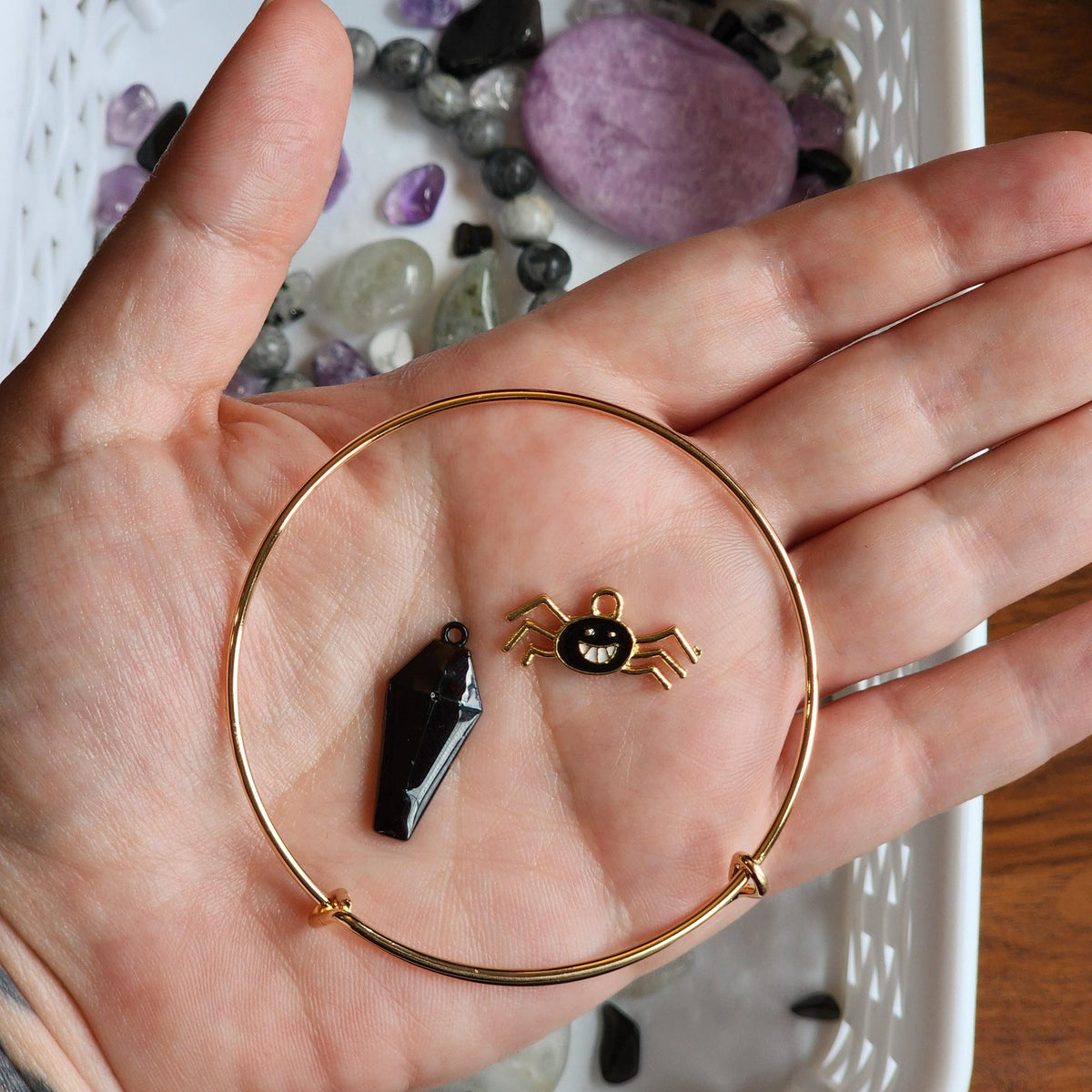 A hand holding a gold bangle with a black spider charm and a purple crystal pendant.