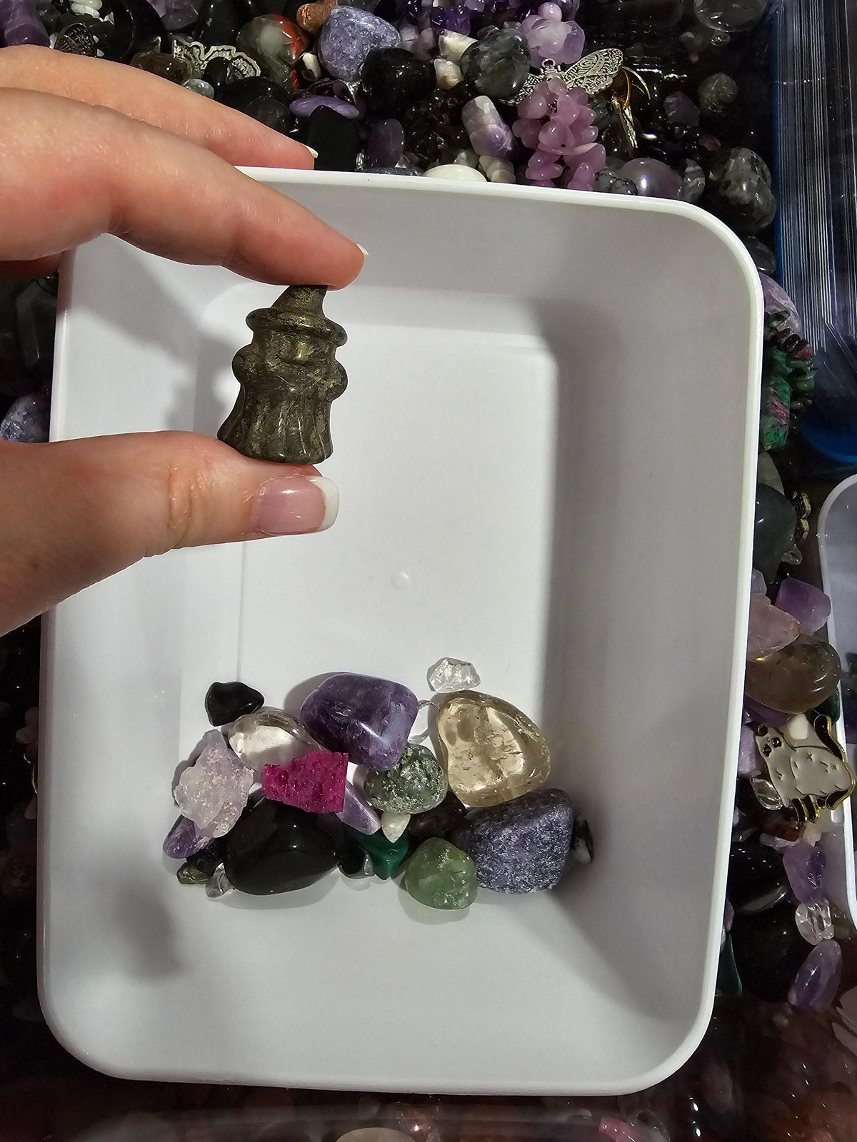 A hand is holding a small figurine above a pile of colorful gemstones in a white container.