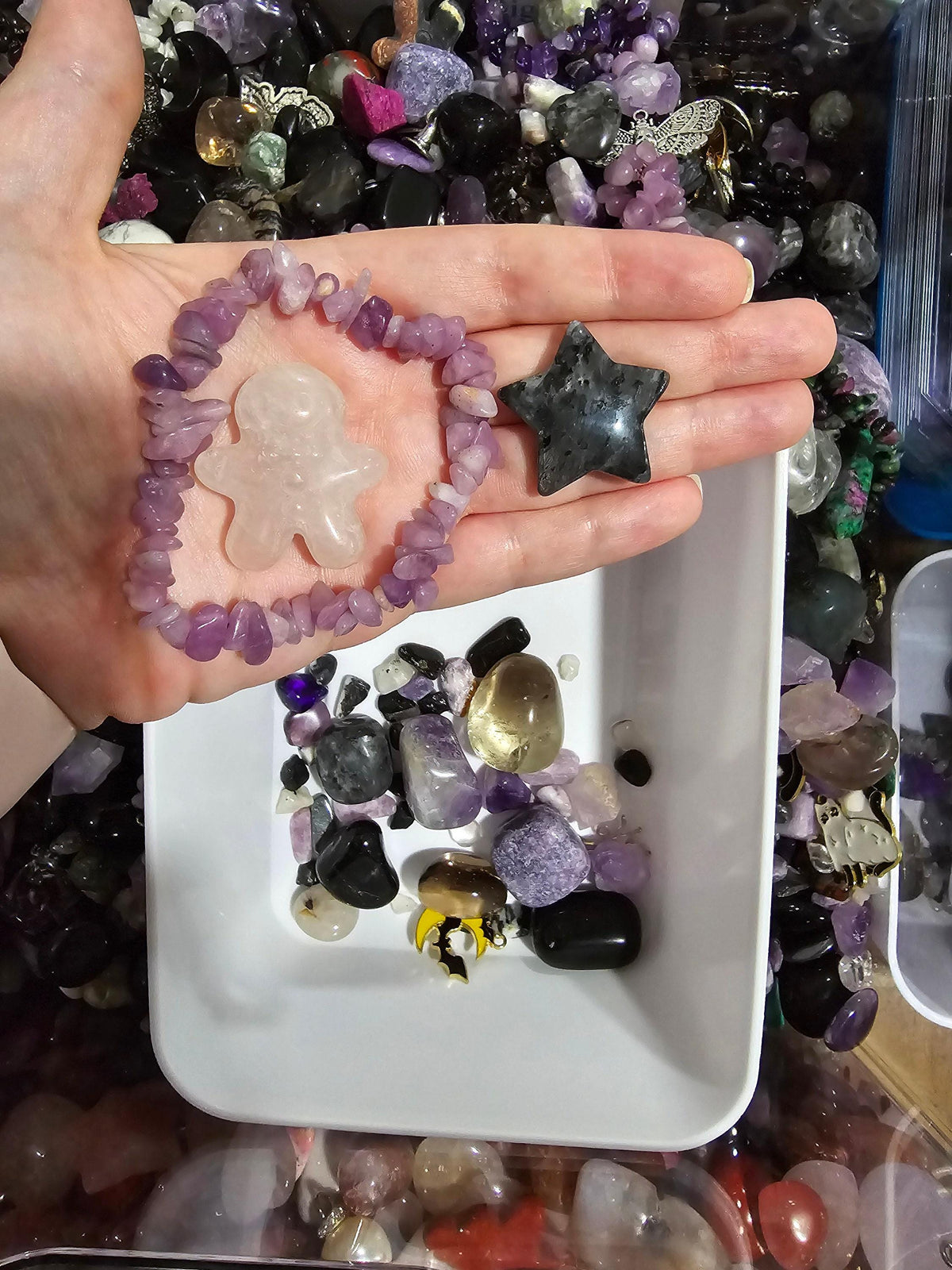 A hand holding a bracelet made of purple stones and a black star-shaped stone, surrounded by various colorful gemstones and crystals in a white bowl.