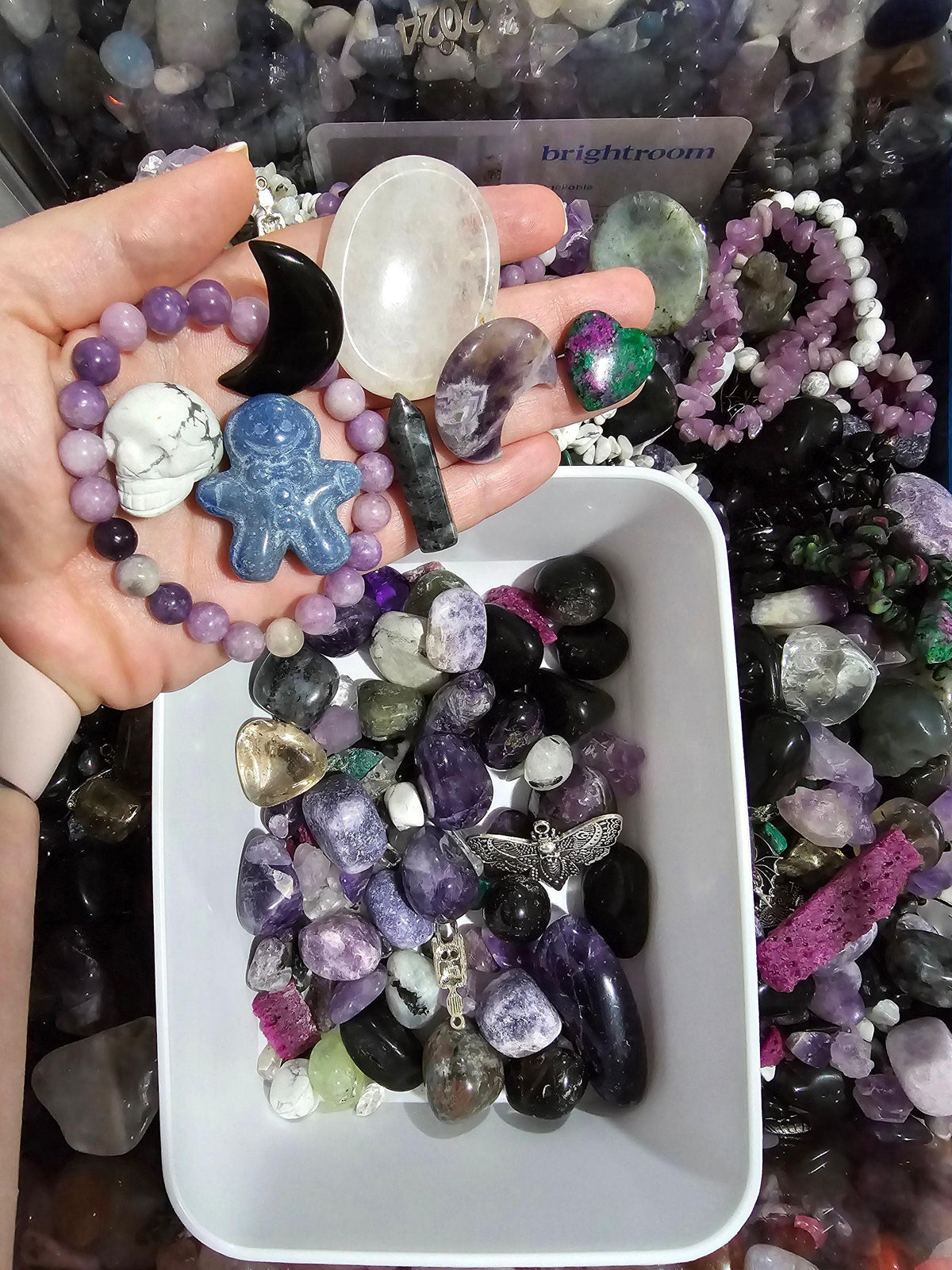 A hand holding various crystals and gemstones, including amethyst, quartz, and amethyst, in front of a display case filled with more crystals and gemstones.