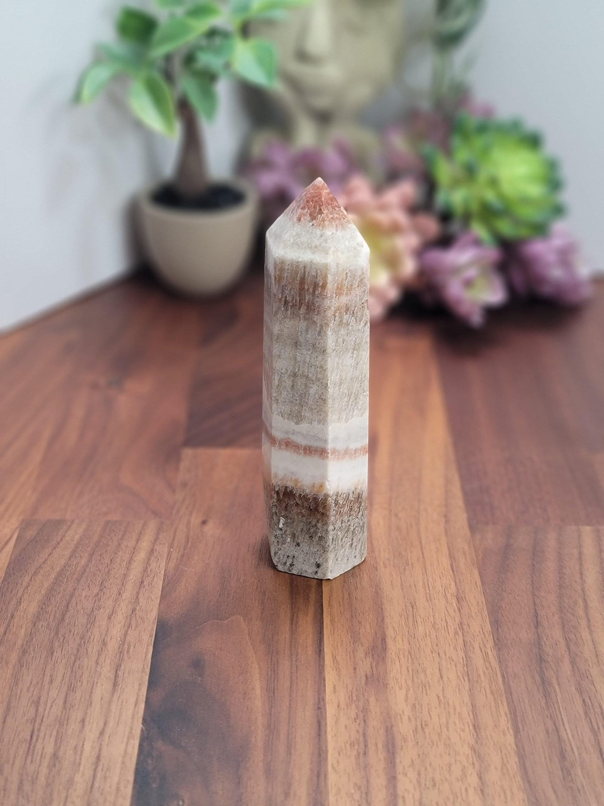 a tall, white crystal or gemstone with a unique striped pattern, placed on a wooden surface. In the background, there is a potted plant and some flowers, creating a natural and serene atmosphere.