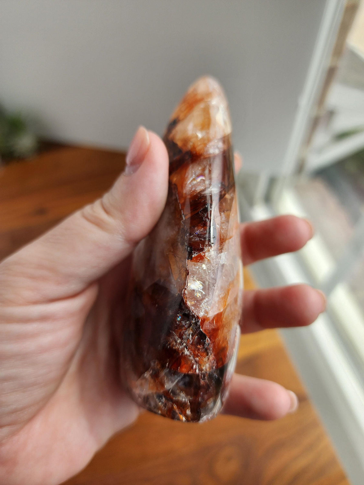 A hand holding a unique, colorful crystal or gemstone, with a blurred background that includes a window and a plant.