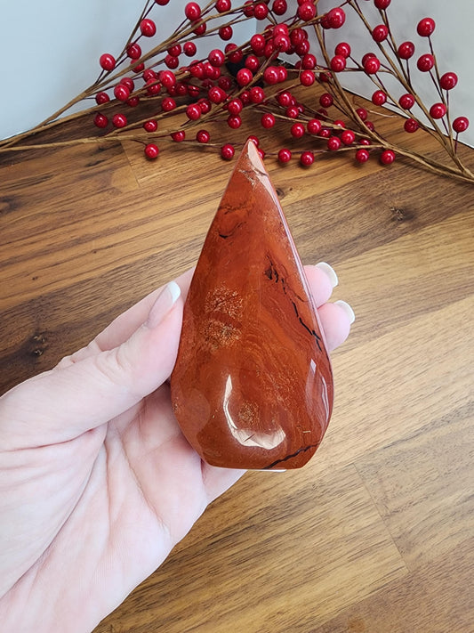 A hand is holding a red, jagged-edged stone on a wooden surface, with a branch of red berries in the background.