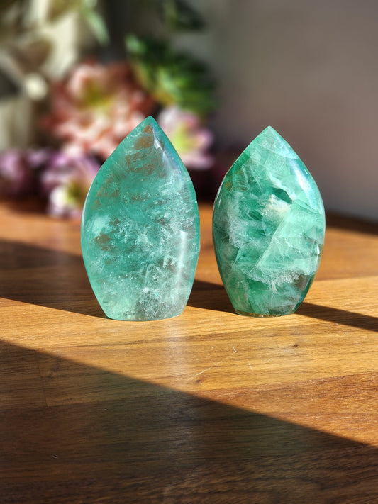 May include: Two green fluorite crystal points, carved into a leaf shape, are displayed on a wooden surface. The crystals are translucent and have a light green color.