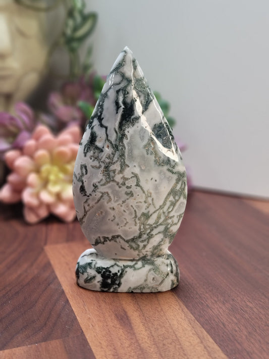 May include: A green and white agate stone carved into a teardrop shape. The stone is polished and has a smooth surface. It is sitting on a wooden surface.