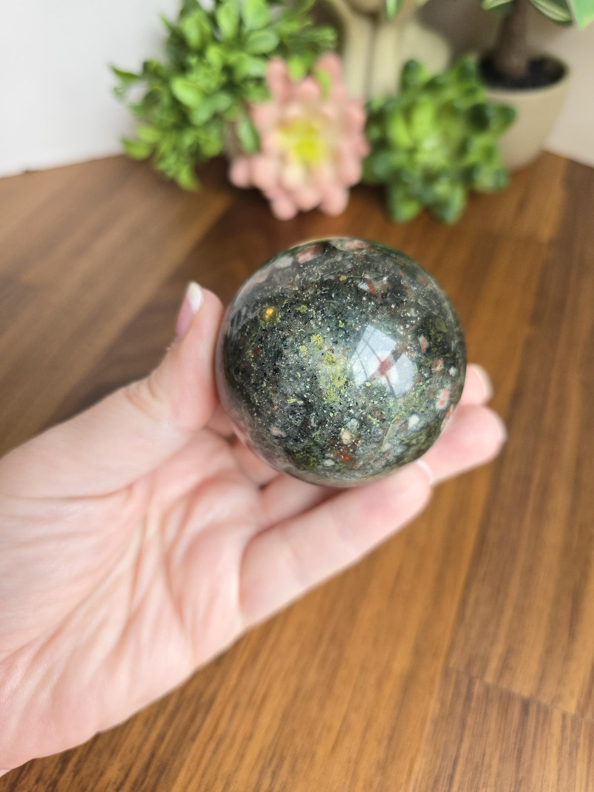 May include: A polished spherical stone featuring a dark green base color with speckled inclusions of red, yellow, and white mineral deposits throughout its surface. The orb has a smooth, reflective texture that catches light across its rounded form.