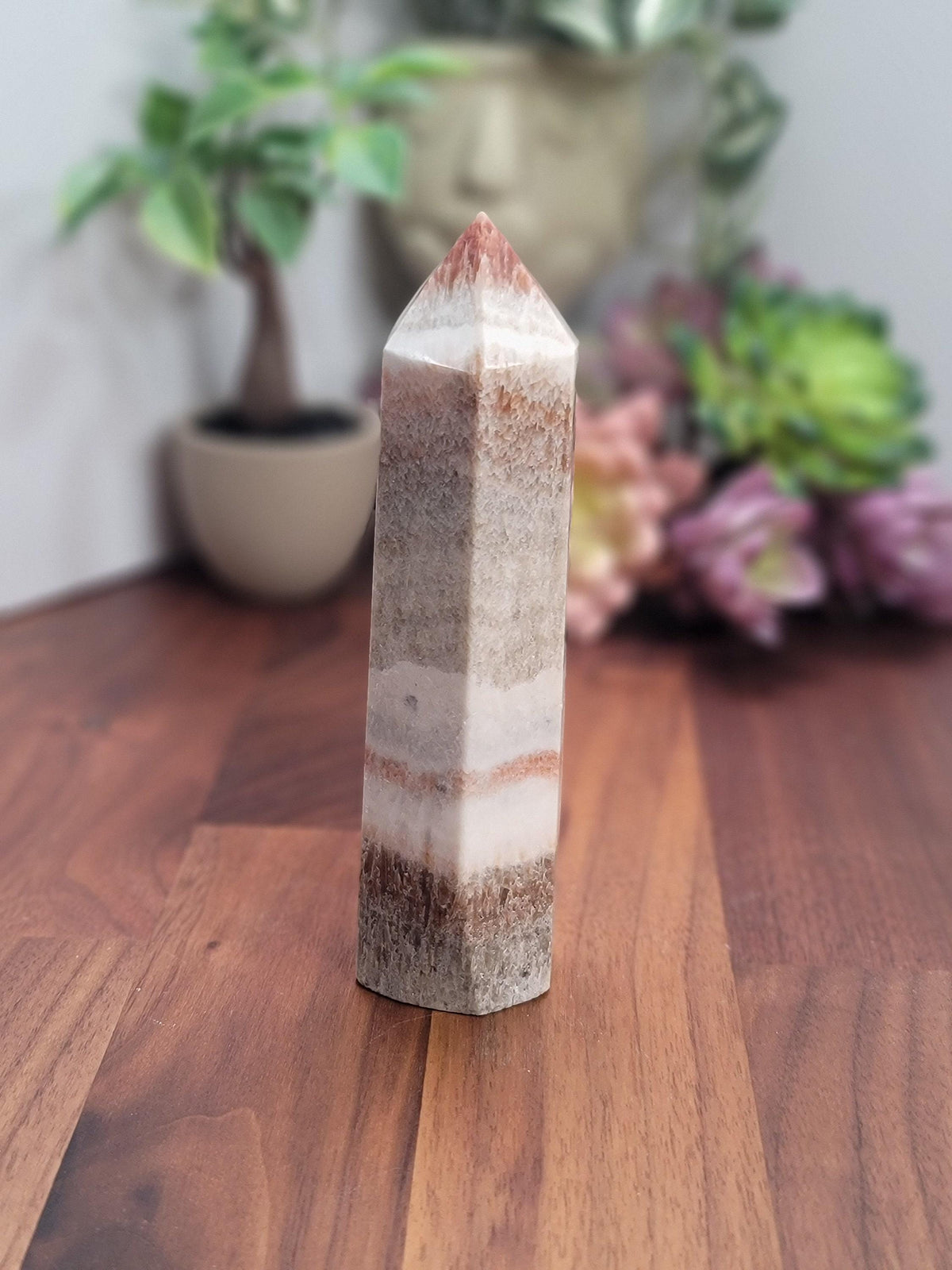 A tall, pointed crystal formation with a striped pattern sits on a wooden surface, with a blurred background featuring a potted plant and flowers.