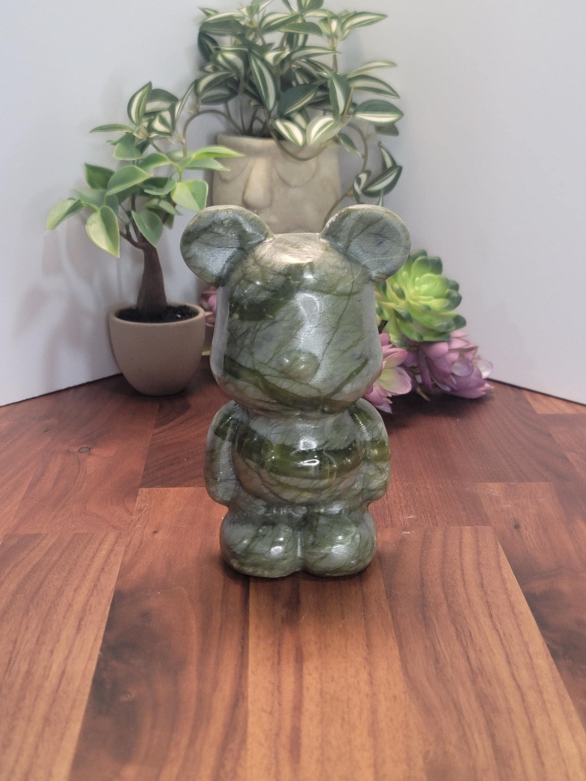 a green stone bear figurine sitting on a wooden surface, surrounded by potted plants and flowers.