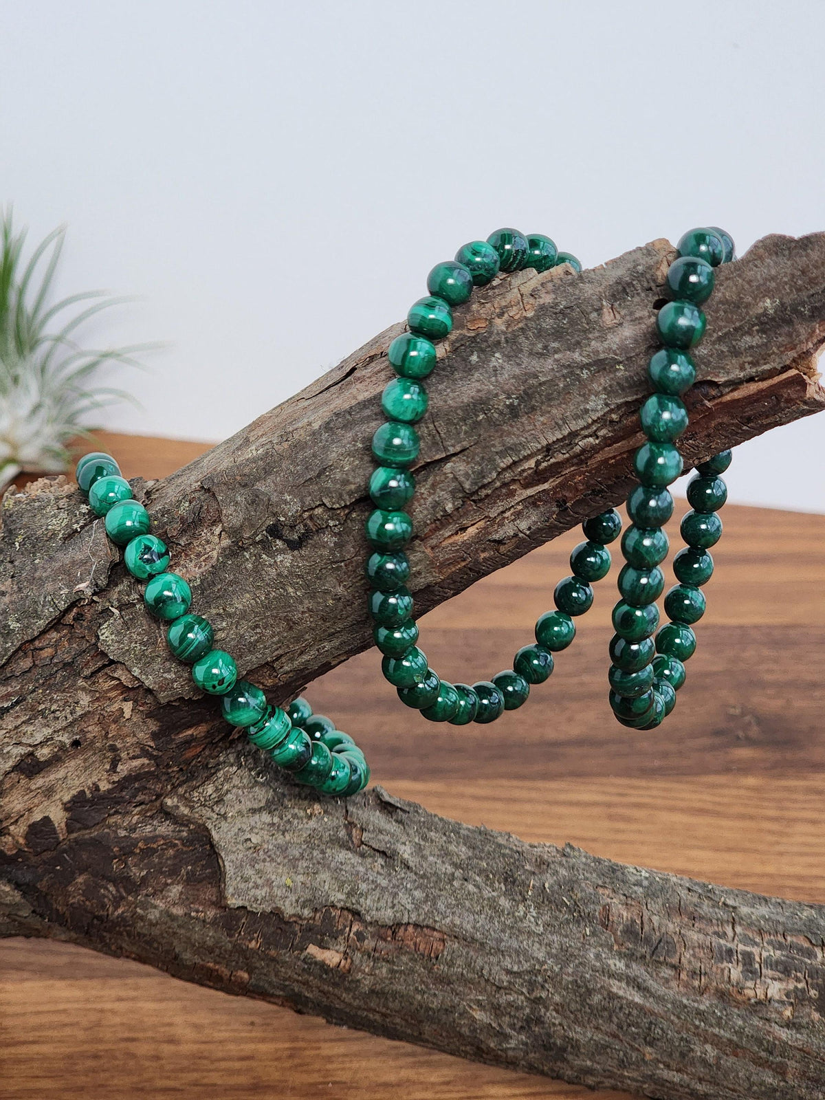 A necklace of green malachite beads is displayed on a wooden surface, with a plant visible in the background.