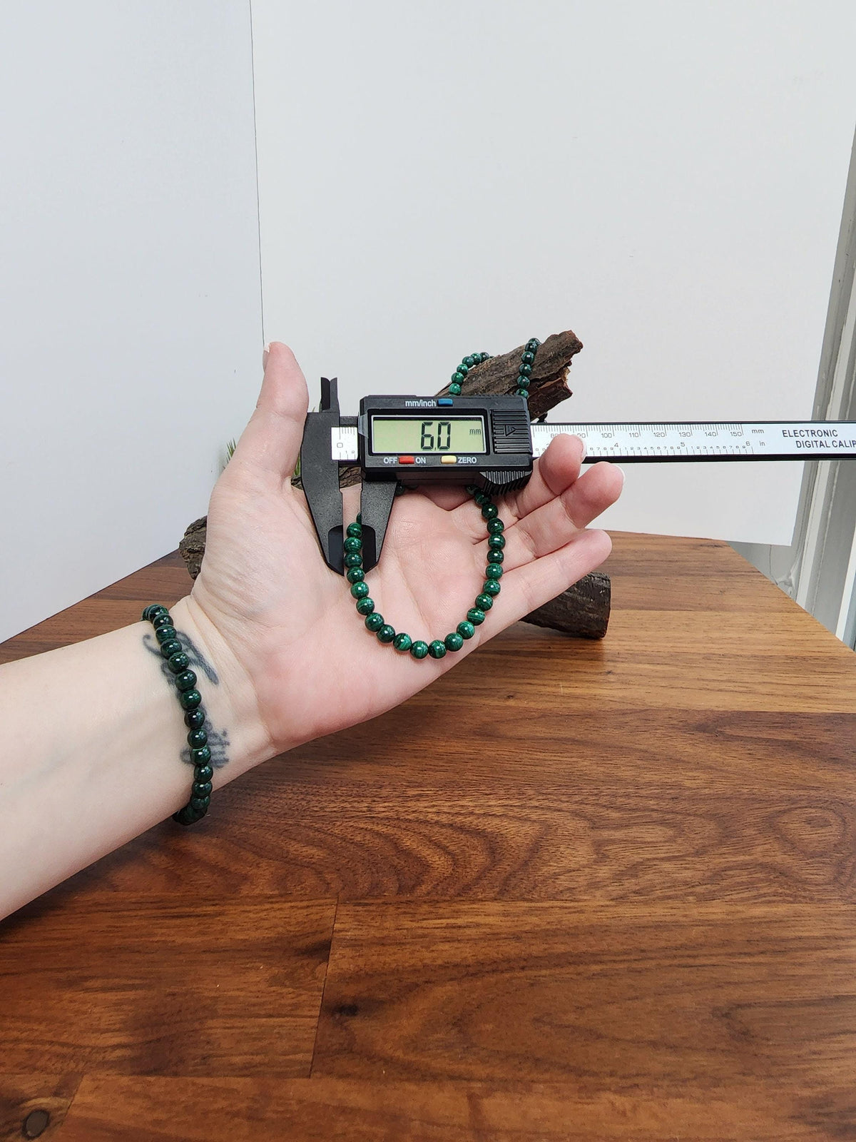 A person's hand holding a digital caliper to measure the size of a green bead bracelet on a wooden surface.
