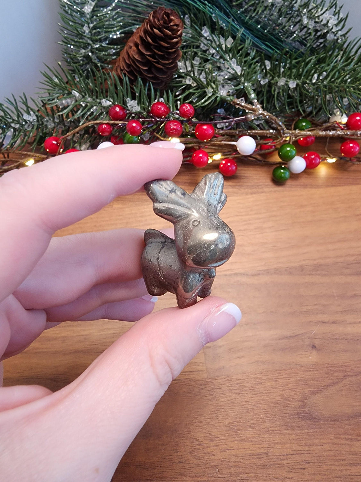 A hand holding a small metal figurine of a reindeer against a backdrop of a Christmas tree with red berries and pine cones.