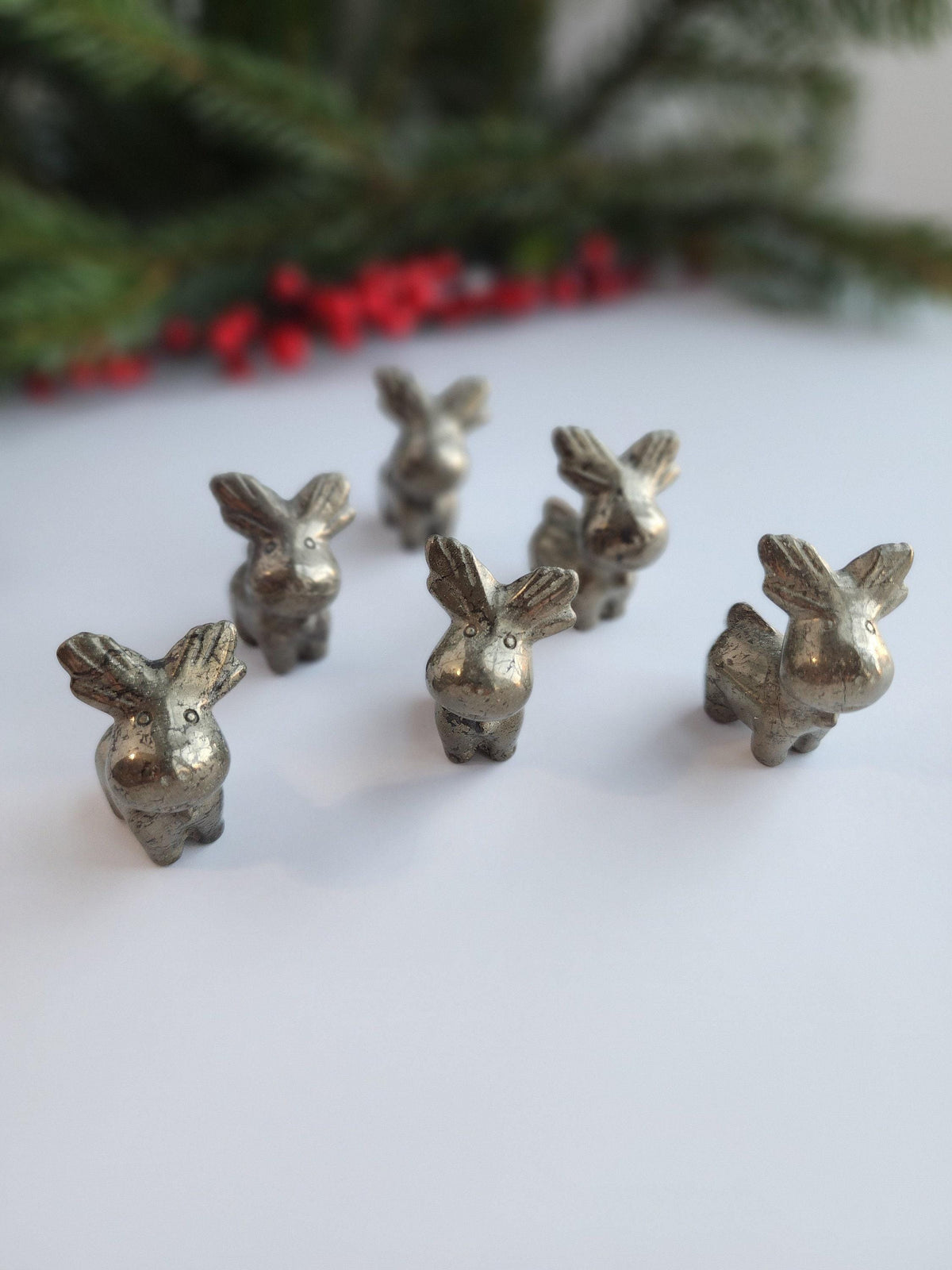 a group of six small, silver-colored metal figurines arranged on a white surface, with a blurred background of greenery and red berries.