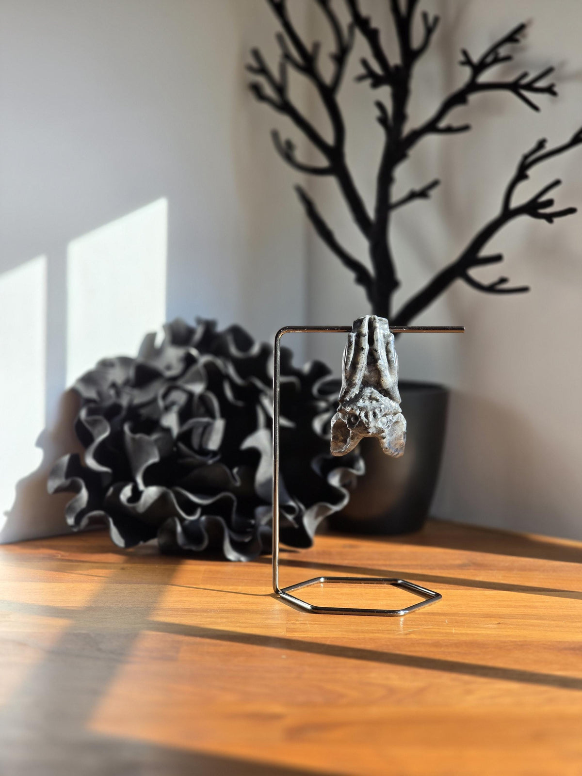 a sculpture of a hand hanging from a metal stand on a wooden surface, with a black tree branch in the background.