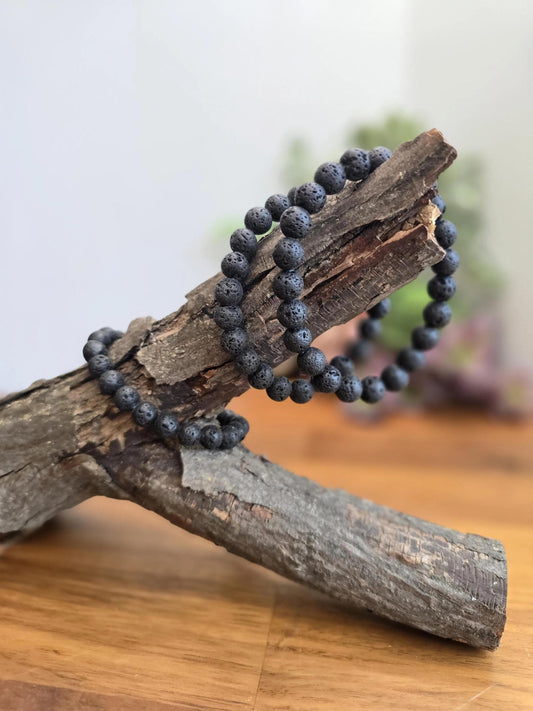 May include: A black lava rock beaded bracelet on a wooden branch. The bracelet is made of small, round beads with a rough, textured surface.