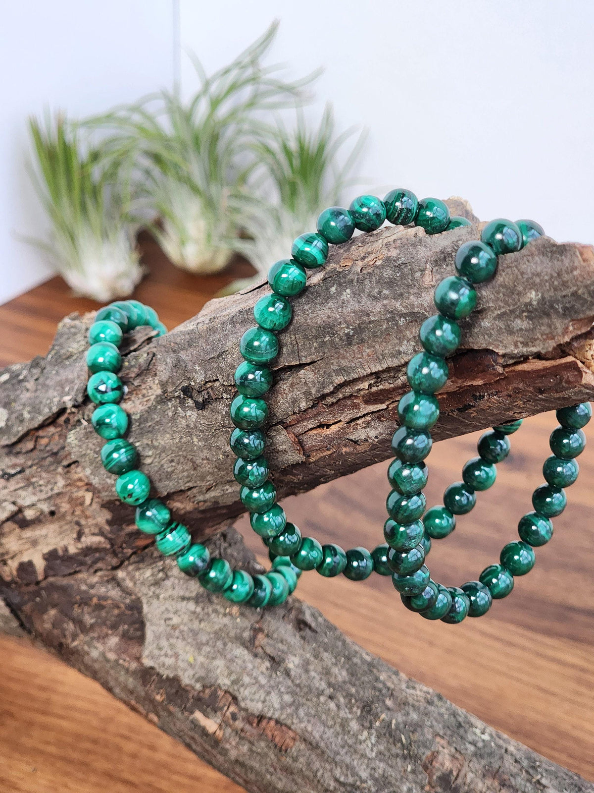 A pair of green beaded bracelets rests on a wooden surface, with a blurred background featuring some plants.